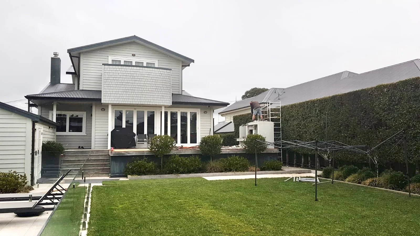 A person installing or repairing a satellite dish on a raised outdoor patio of a gray house with white trim. The yard has green grass and a hedge.