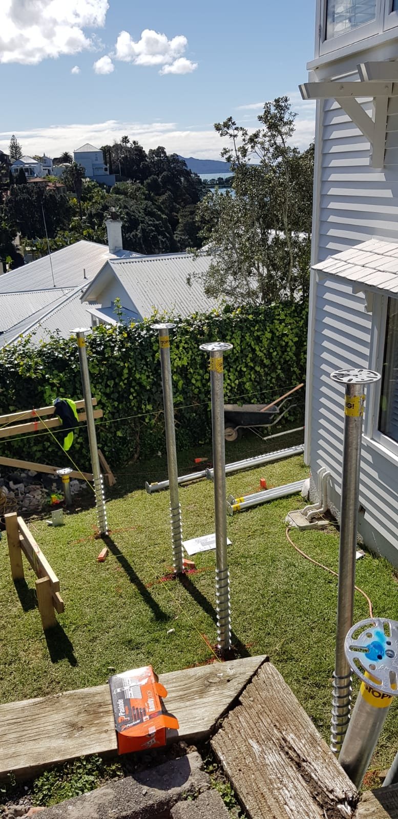 A backyard under construction with steel supports for a deck or roof, a wheelbarrow, and orange string guides. The scene overlooks neighboring houses, trees, and a body of water in the distance.