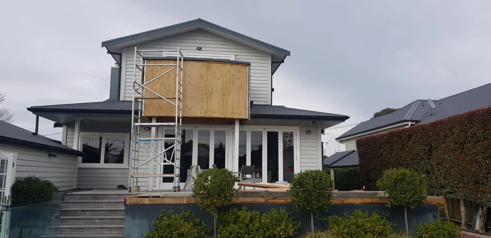 A two-story house undergoing renovation with a large wooden panel on the upper exterior wall, a scaffolding structure, and a deck with gardening tools in front.