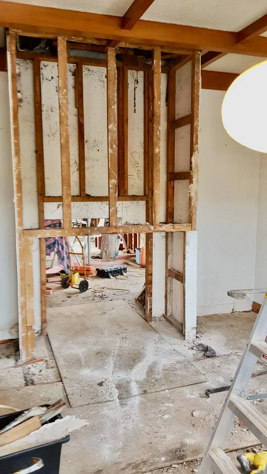 View of a wall under renovation, showing exposed wooden studs and partial drywall. Construction tools and debris are scattered on the unfinished floor.