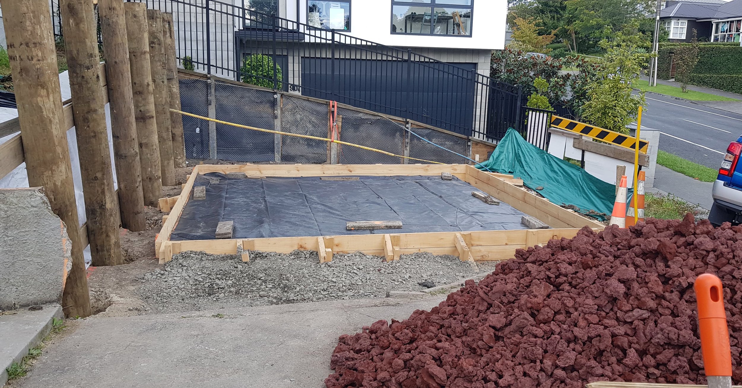 Construction site with a wooden frame and black plastic-covered foundation, orange safety cones, large mound of reddish gravel, and various construction materials, with fenced-in yard and residential street in the background.