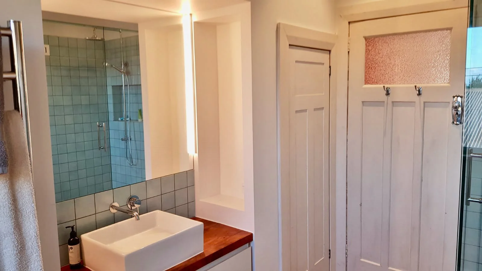 A modern bathroom with a white rectangular sink, wooden countertop, a mirror, a glass-enclosed shower area with blue tiles, and a wooden door.