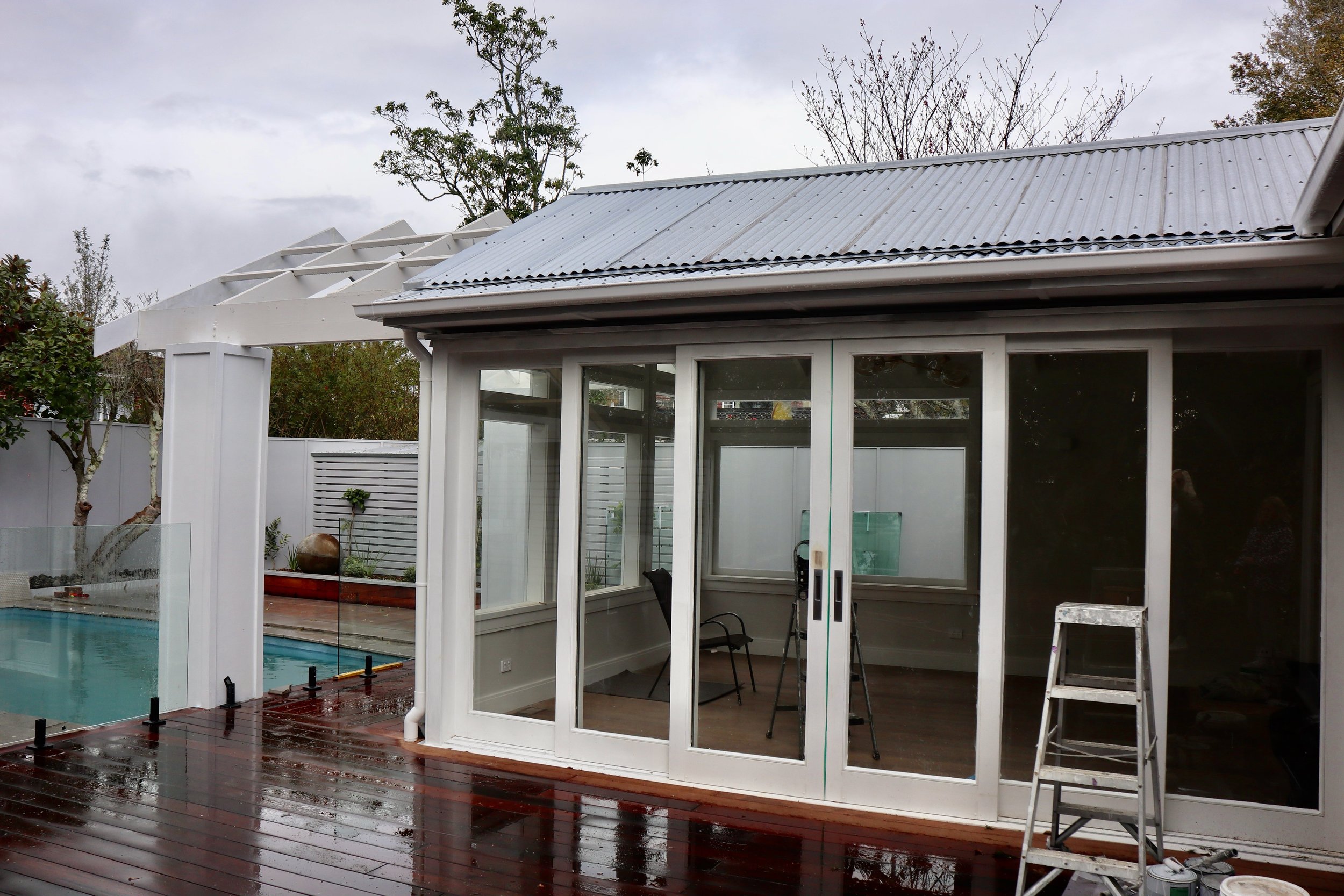 A newly renovated backyard with a swimming pool, a glass sunroom with sliding doors, wooden deck, and ongoing construction work visible with a ladder and construction tools.