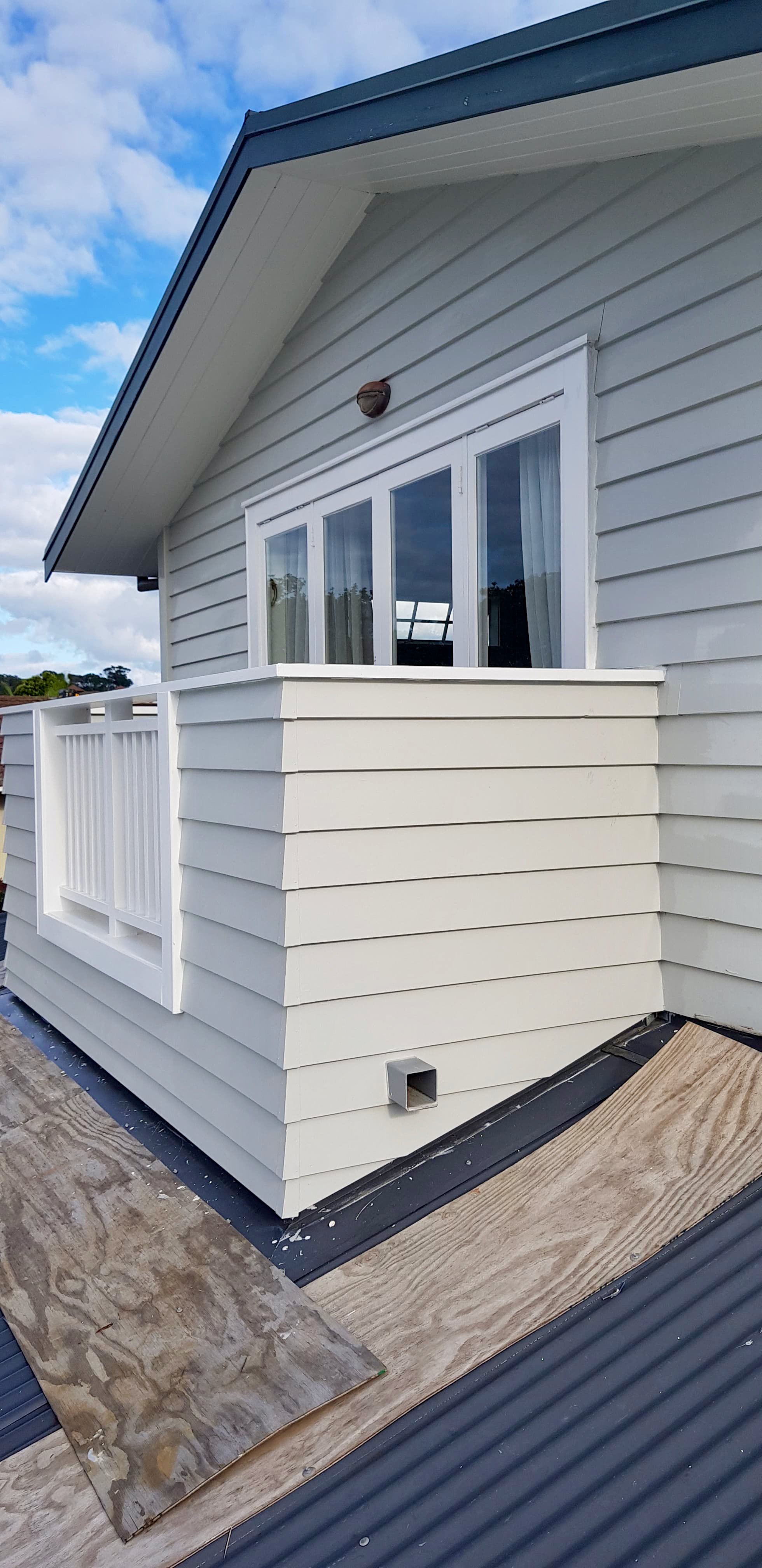 A view of a house exterior with light gray siding, a small balcony with a white railing, and a window with white frames. Part of the roof with black shingles and a small dry vent pipe are visible. The sky is partly cloudy.