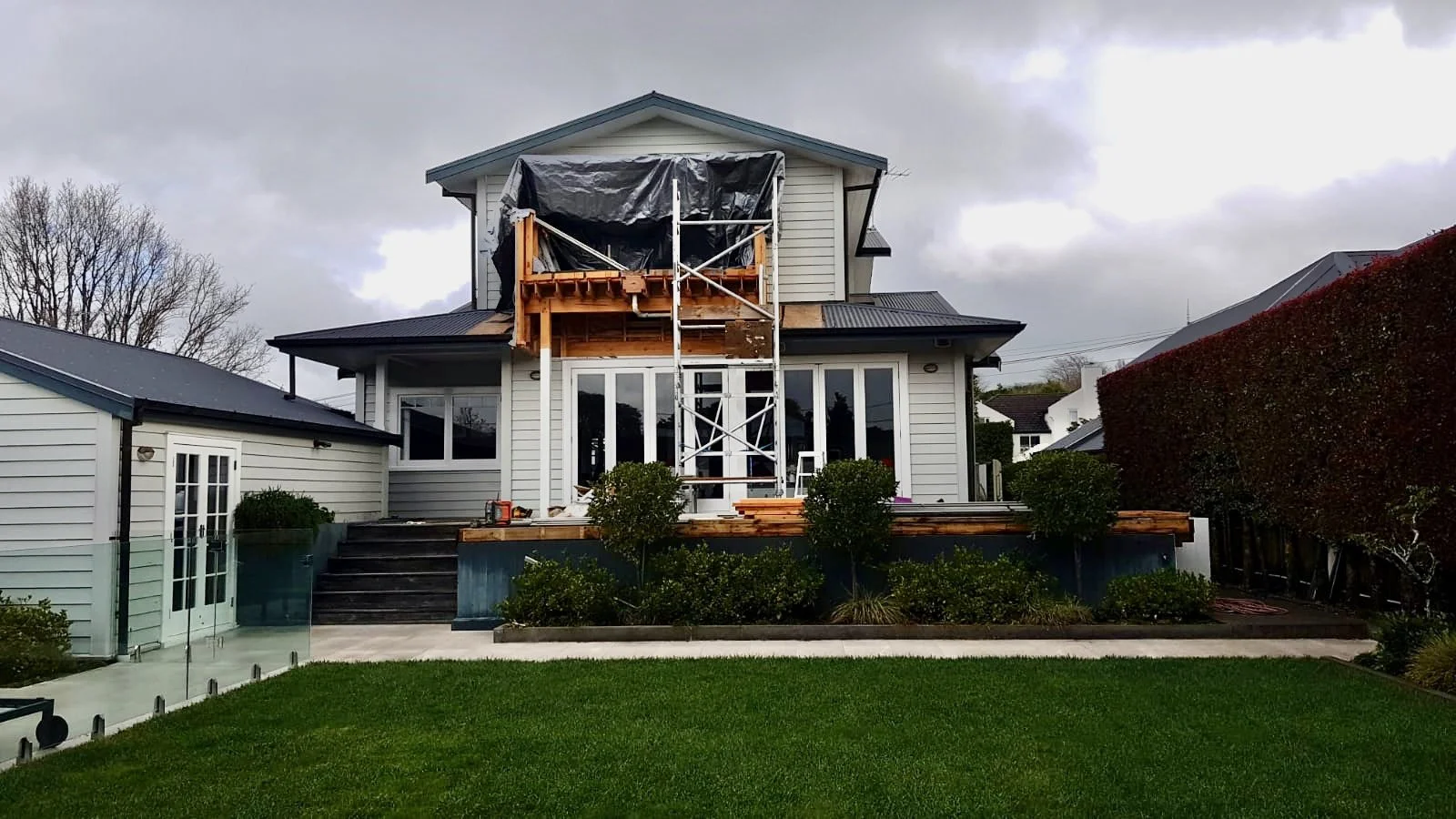 A house under construction with scaffolding in front of an open balcony area. The house has white siding, large glass doors, and a dark roof. The front yard features a well-kept green lawn and bushes.