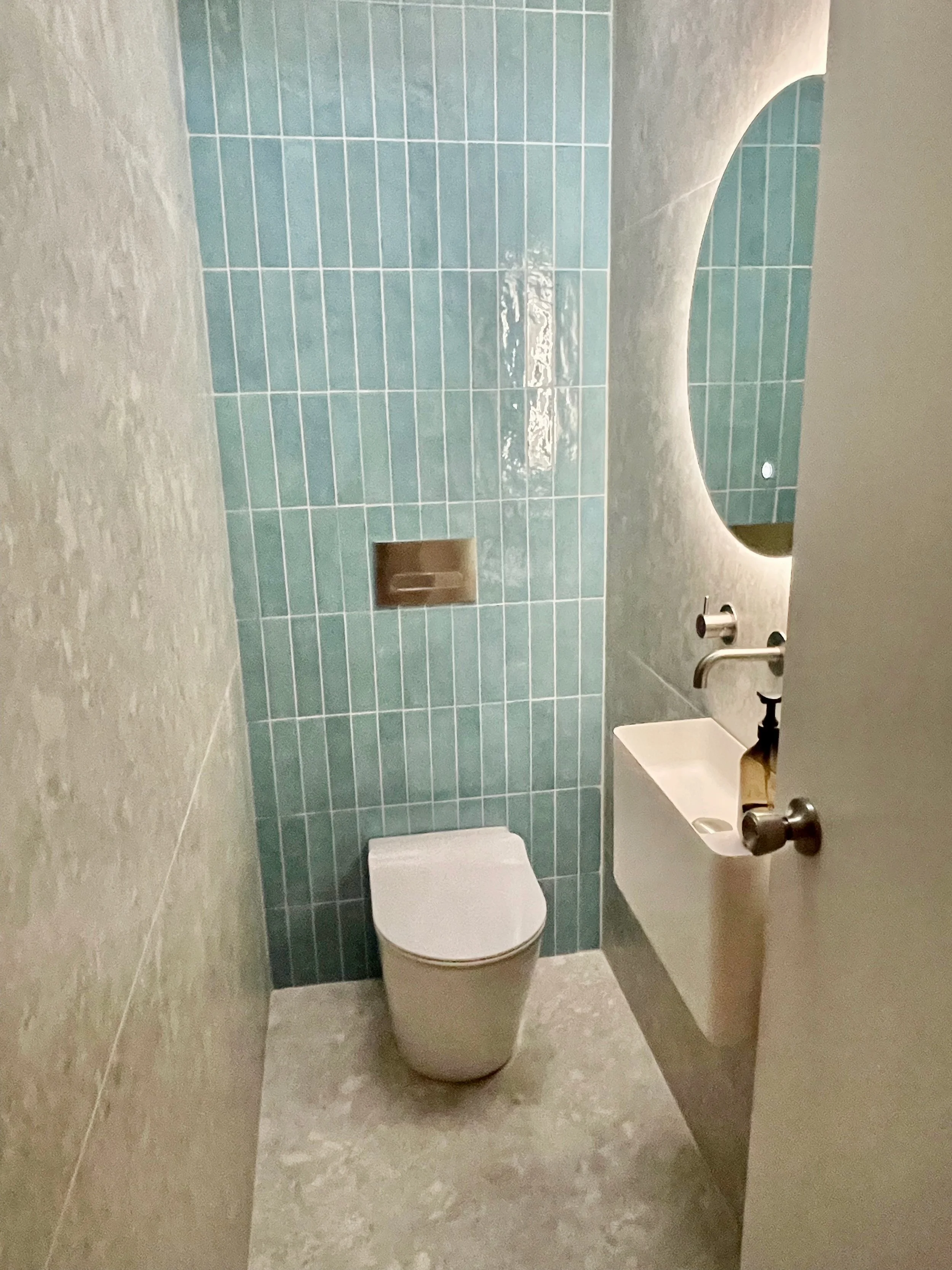 Small modern bathroom with a white toilet, a small white sink, a large oval mirror, and blue tiled wall behind the toilet.