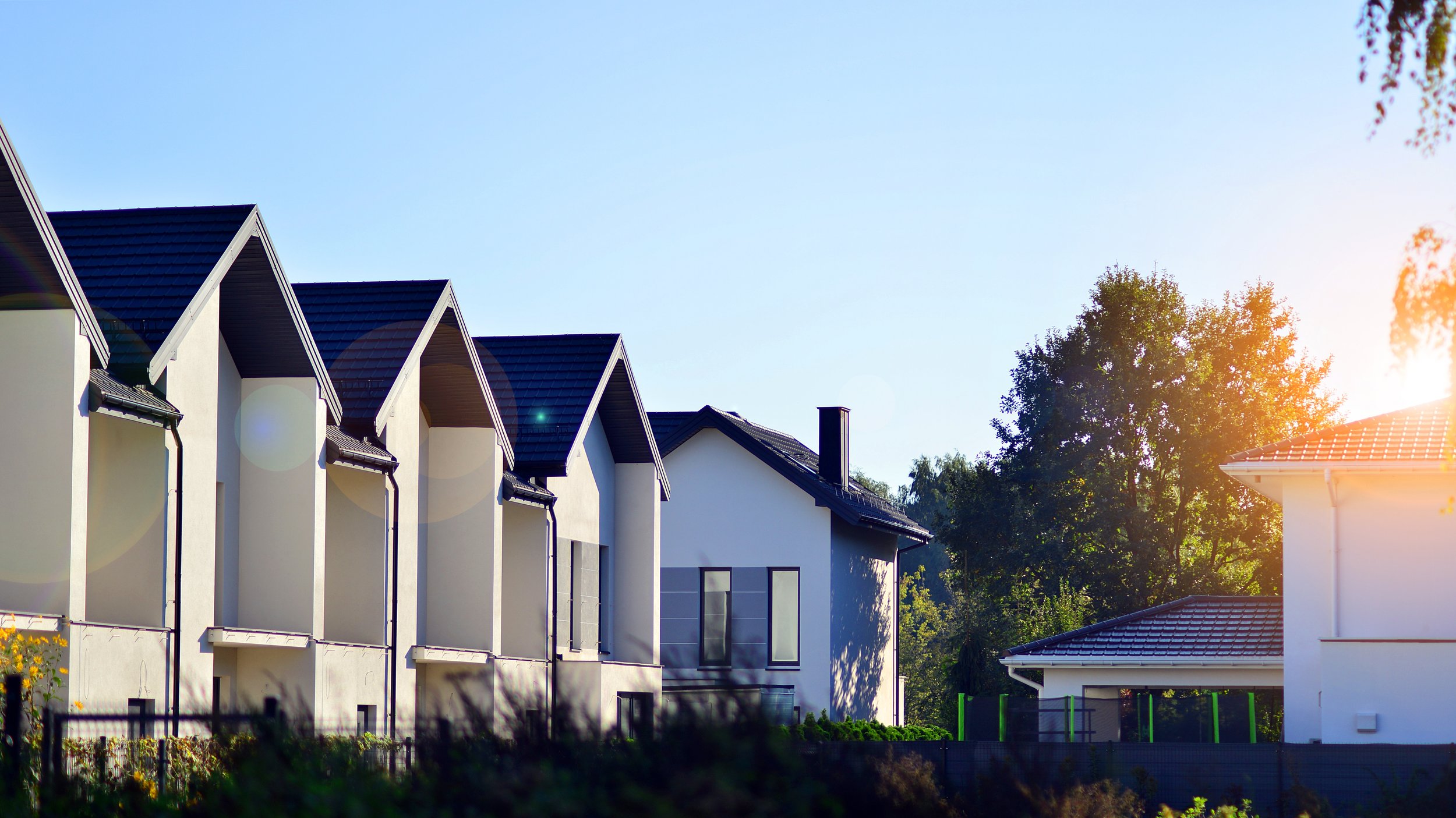 A row of modern white houses with dark roofs, set against a sunny sky and surrounded by trees.