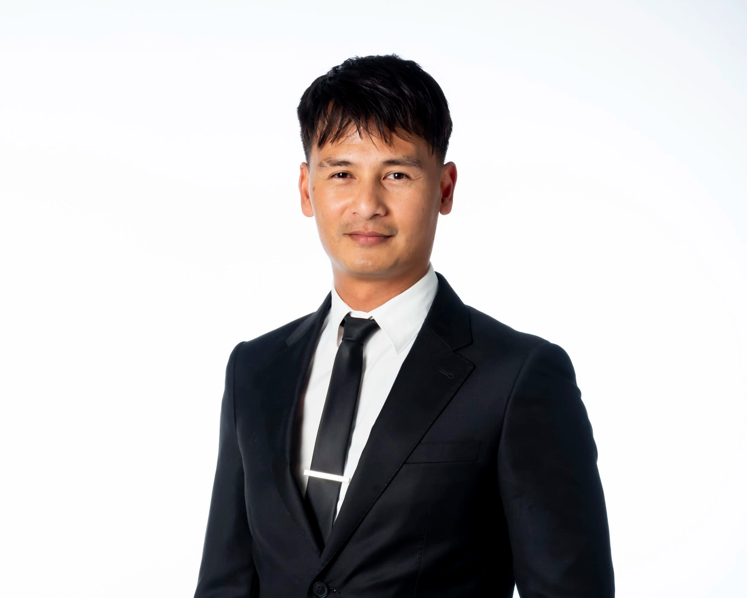 A young man in a black suit, white shirt, and black tie, standing against a plain white background.
