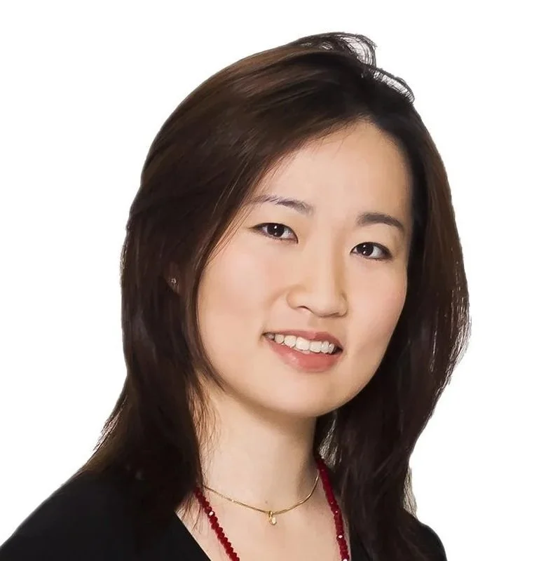 A woman with shoulder-length dark brown hair, wearing a black top, a small gold necklace with a pendant, and a red beaded necklace, smiling slightly against a plain white background.