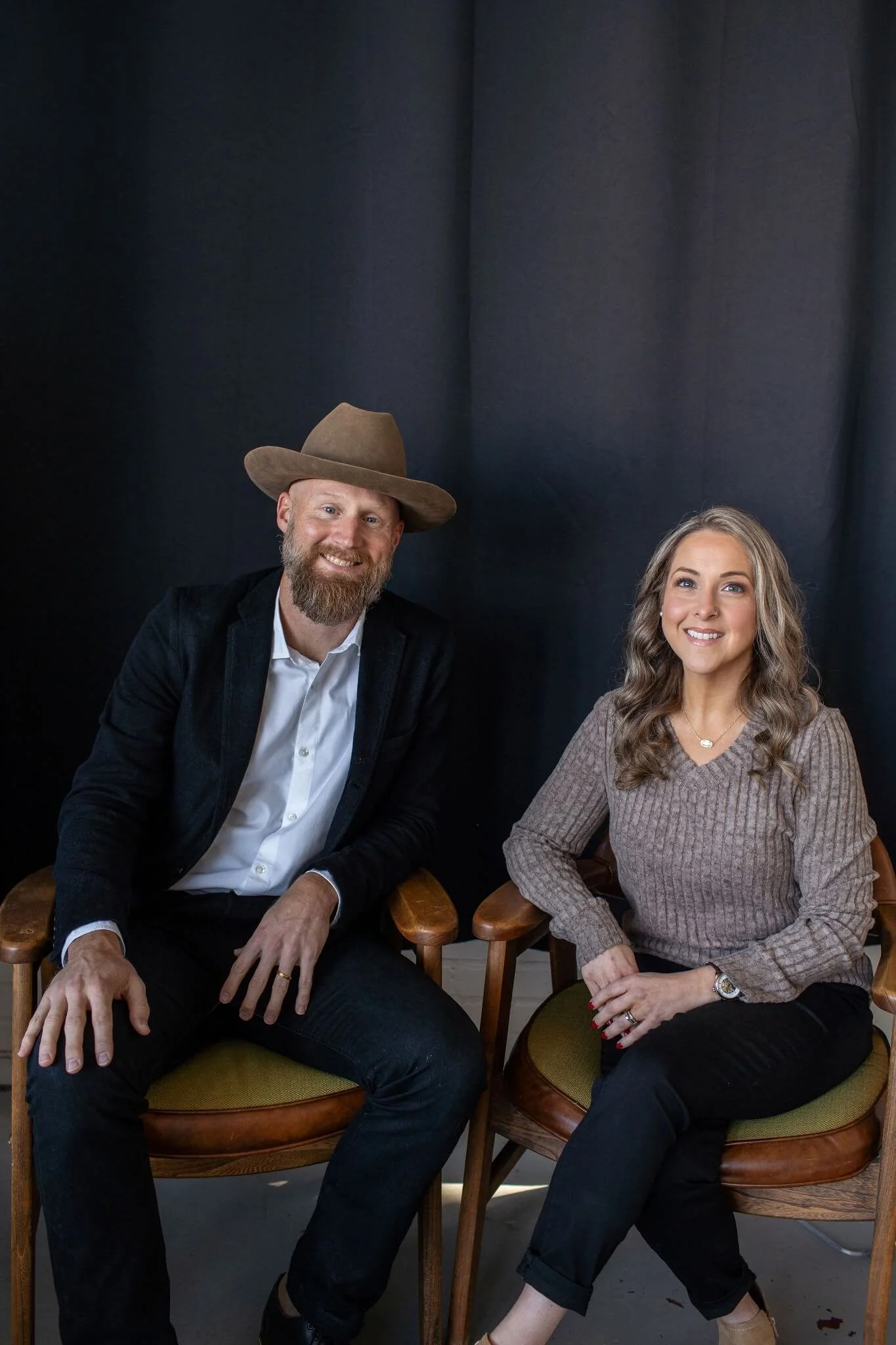 A man with a beard and light skin, wearing a cowboy hat, white shirt, and dark jacket, sitting on a wooden chair. A woman with long curly hair, light skin, wearing a brown sweater and black pants, sitting on a similar chair. Both are smiling against a dark backdrop.