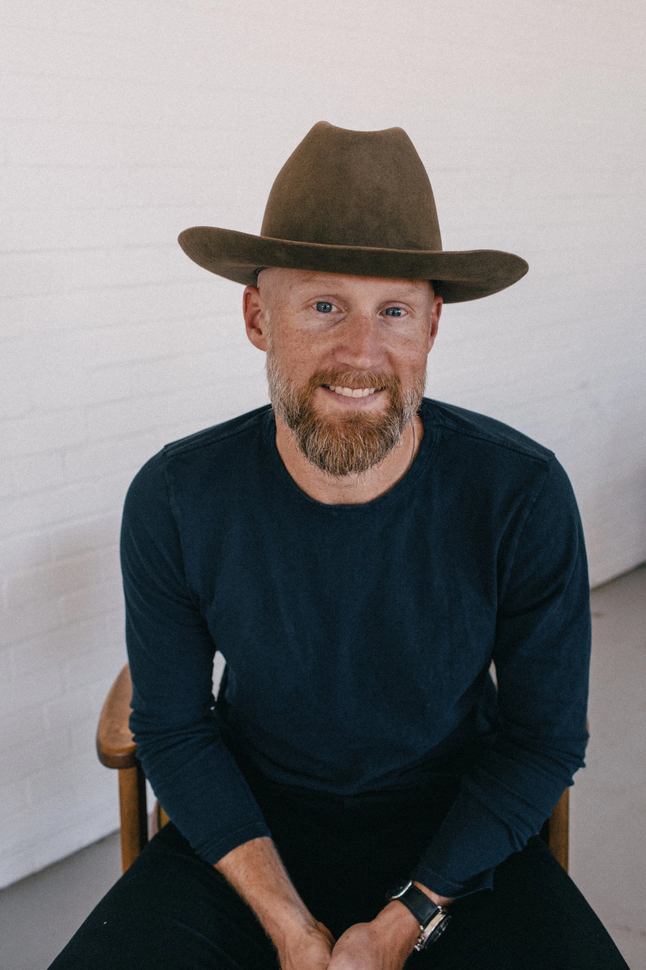 A smiling man with a beard and blue eyes, wearing a wide-brimmed brown hat and a dark long-sleeve shirt, sitting on a wooden chair against a white brick wall.
