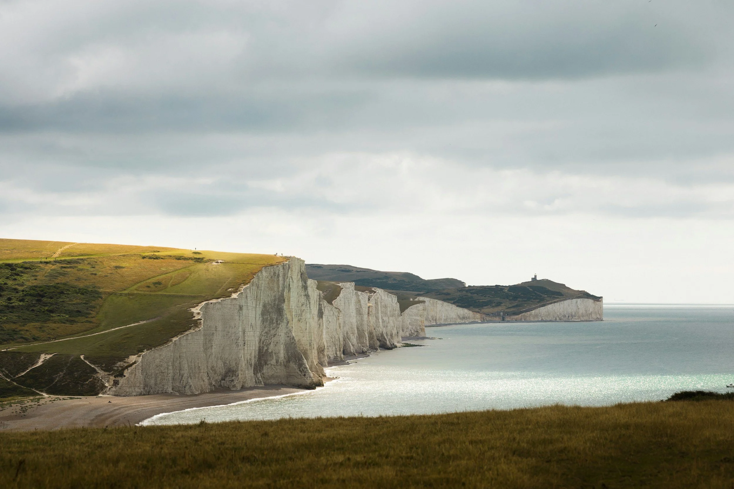 Cliffs of Dover coastal landscape with white chalk cliffs, green grassy areas, and a lighthouse in the distance under a cloudy sky.