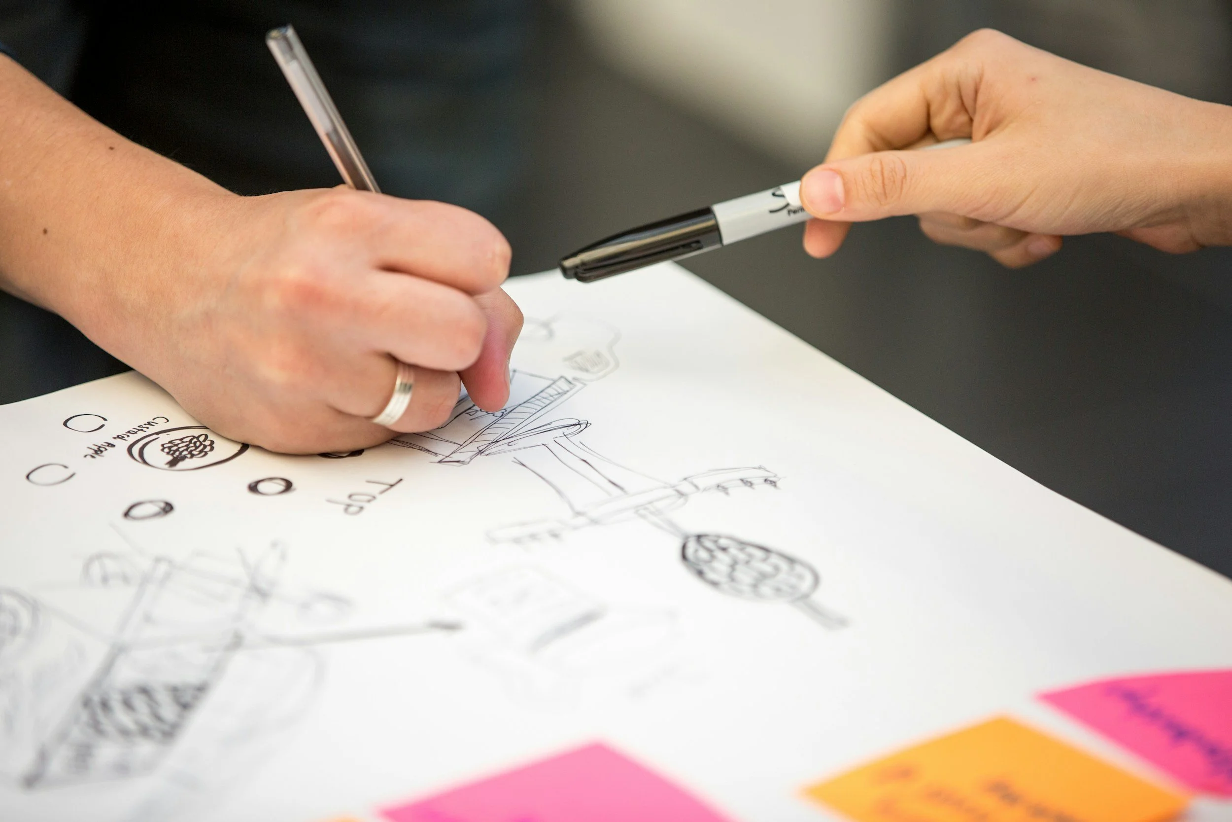 Two people collaborating on a sketch or diagram on a sheet of paper, one person drawing with a pen and the other holding a marker.