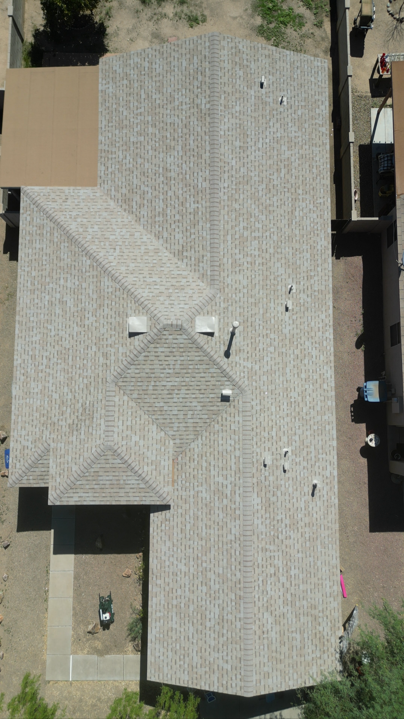 Aerial view of a house with a multi-segmented shingle roof, including gables and vents, and surrounding yard with a concrete walkway and some outdoor furniture.
