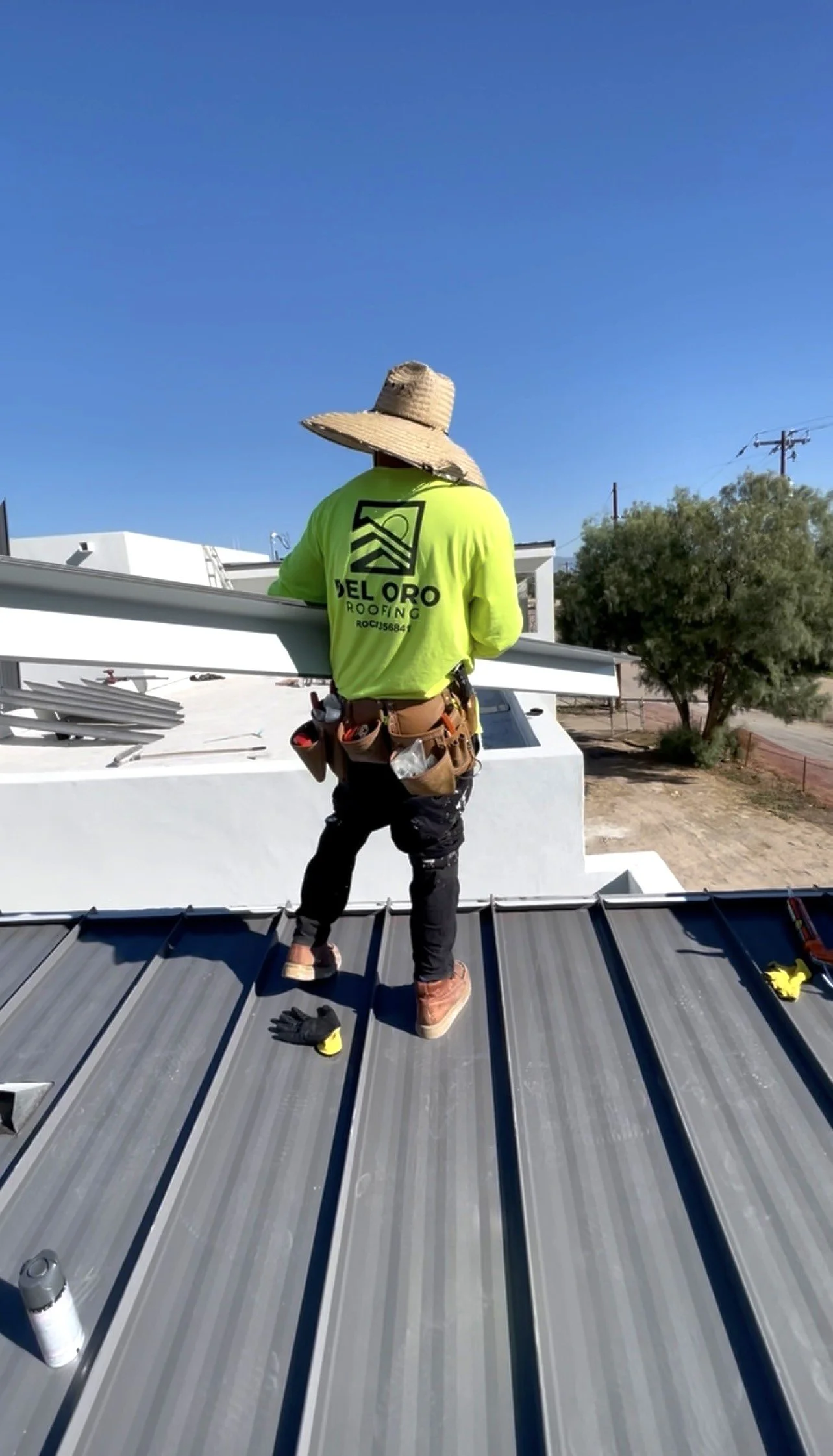 A roofer wearing a neon yellow shirt, jeans, and a wide-brimmed hat working on a metal roof on a sunny day.