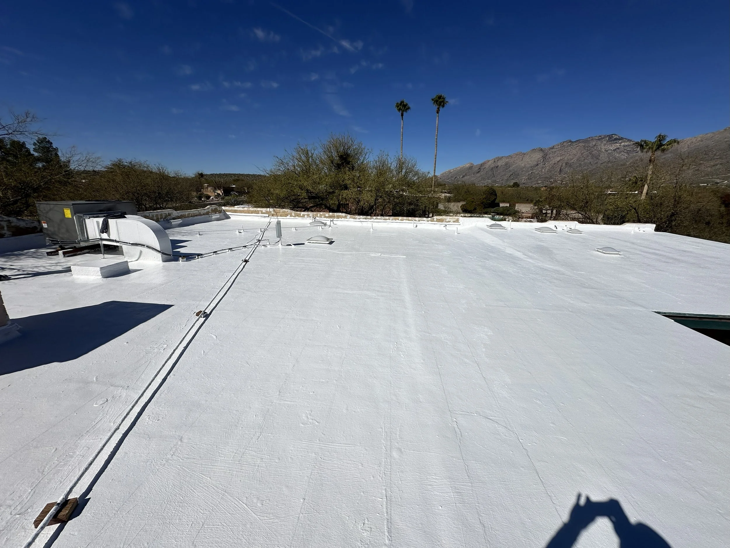 White flat roof with HVAC units, vents, and electrical wiring, with a desert landscape and mountains in the background under a blue sky.