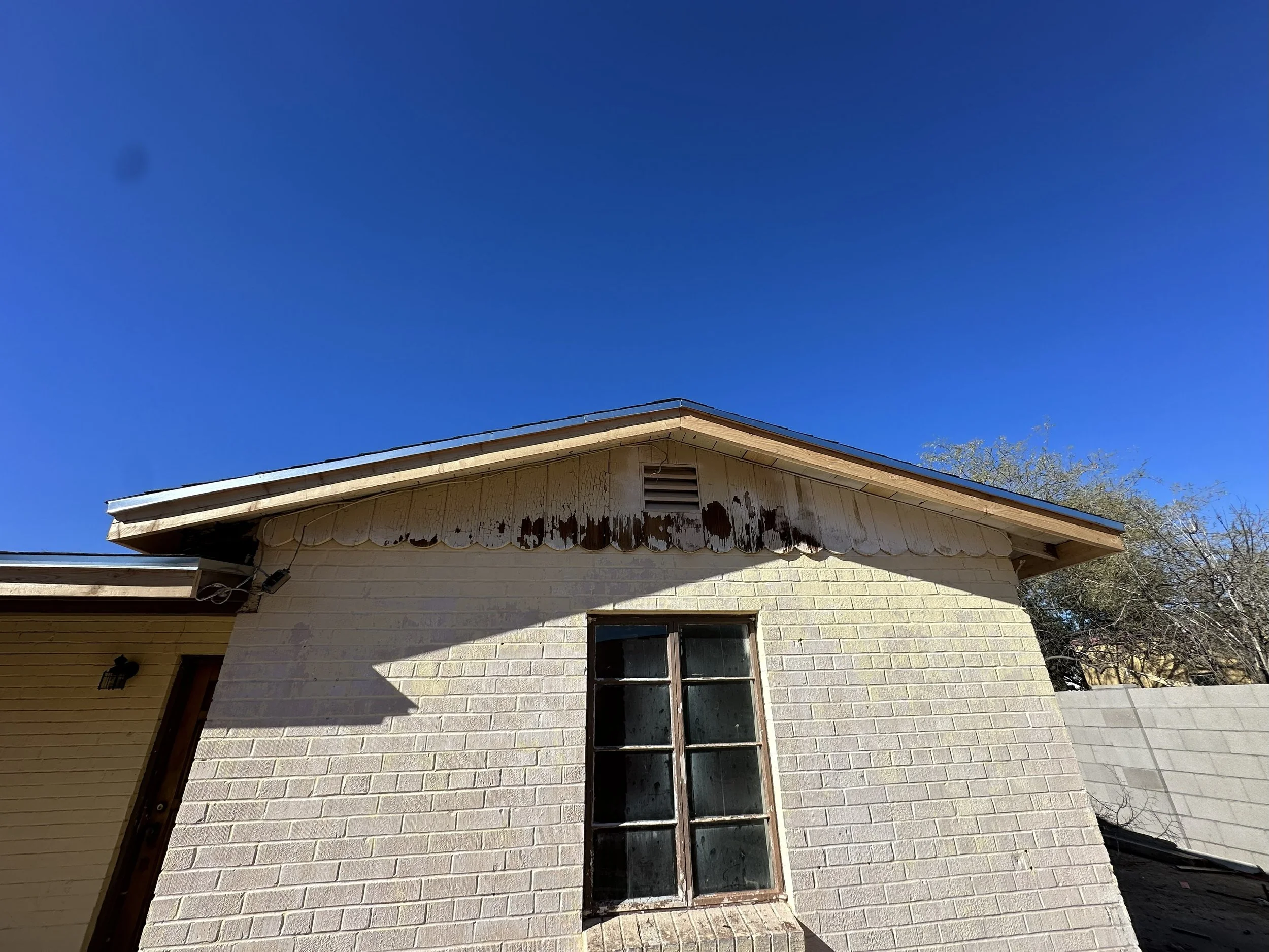 Close-up of a house's exterior wall with a window, showing peeling paint and weathering on the eaves under a clear blue sky.