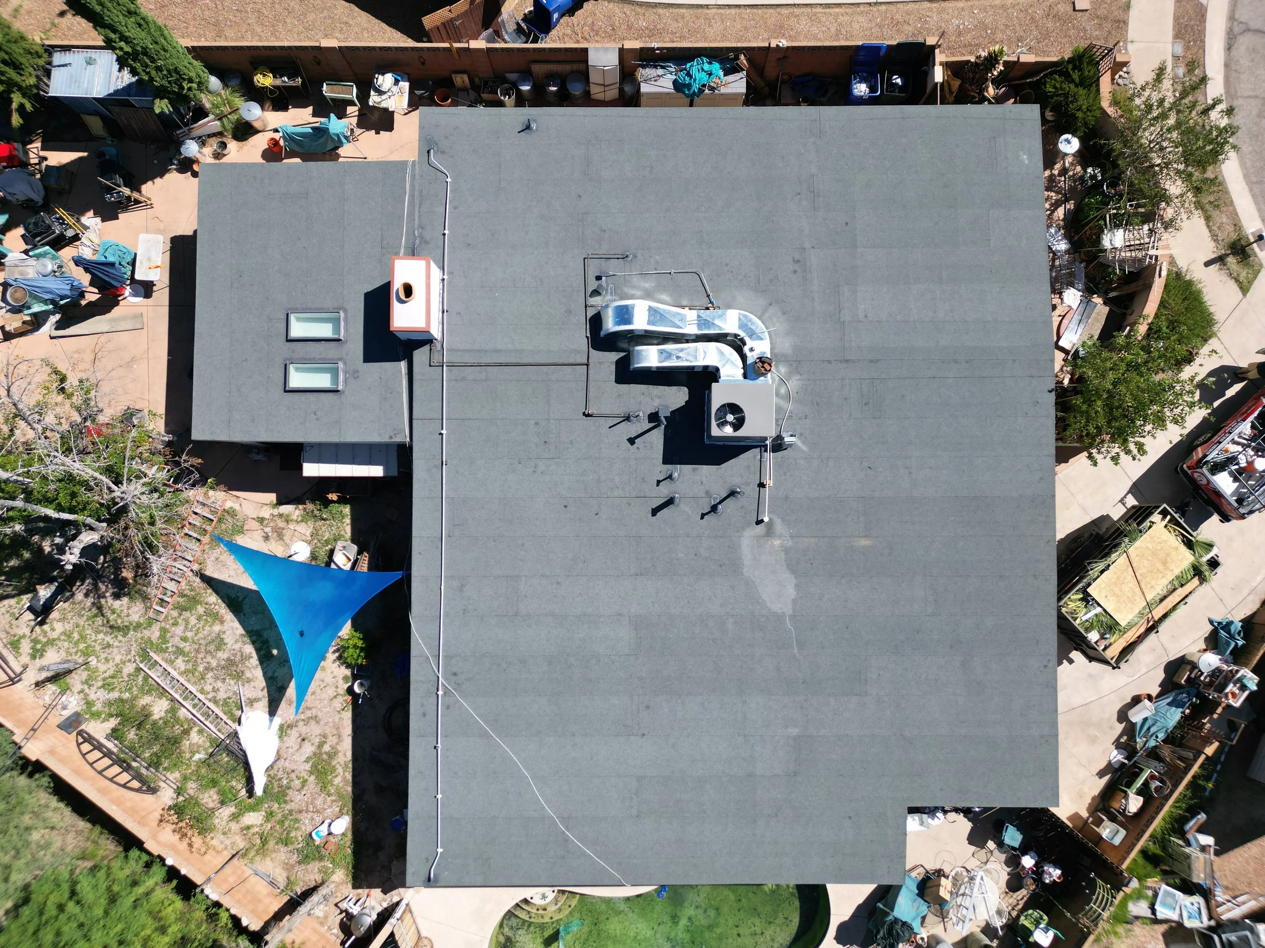 Aerial view of a residential backyard with a flat gray roof, rooftop ventilation units, a small set of skylights, a blue shade sail over a garden bed, and surrounding outdoor furniture and objects.