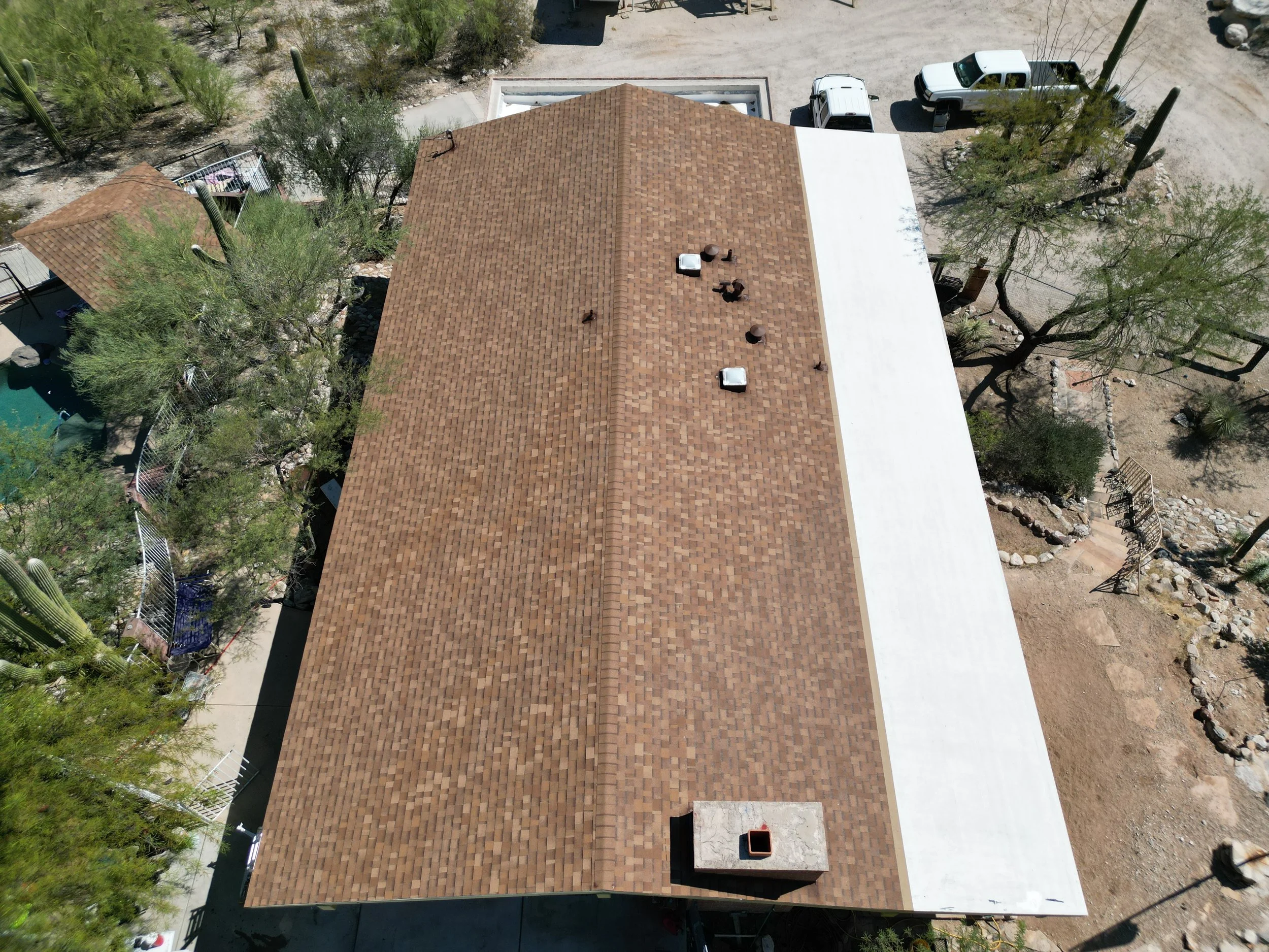 Aerial view of a house with a brown shingle roof, surrounded by desert landscaping with cacti and trees, and a white section of roof on one side.