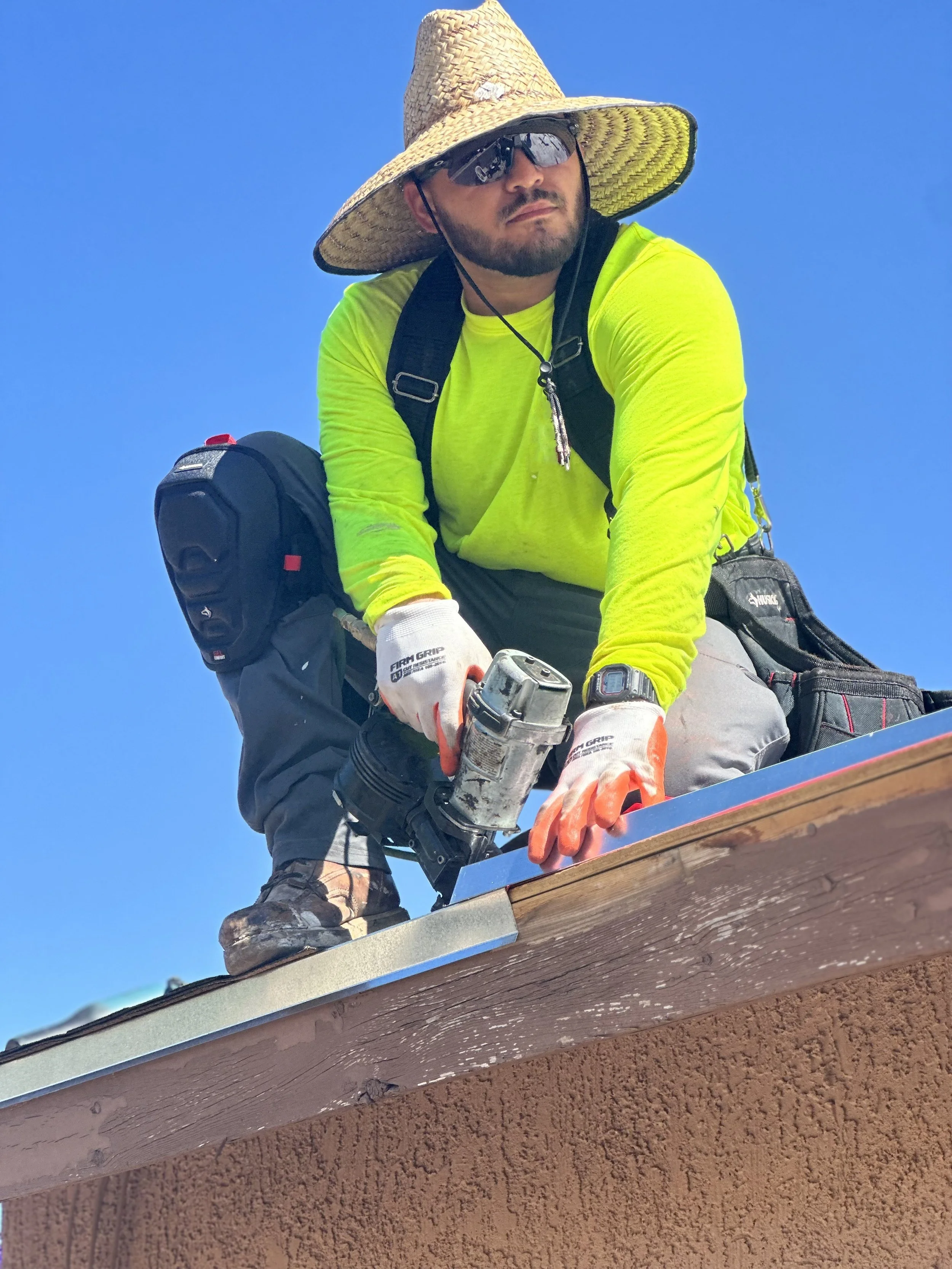 A man wearing a straw hat, sunglasses, and gloves is kneeling on a roof, using a power tool to work on the shingles. He is dressed in a bright yellow shirt and has a backpack and other gear around him. The sky is clear and blue.