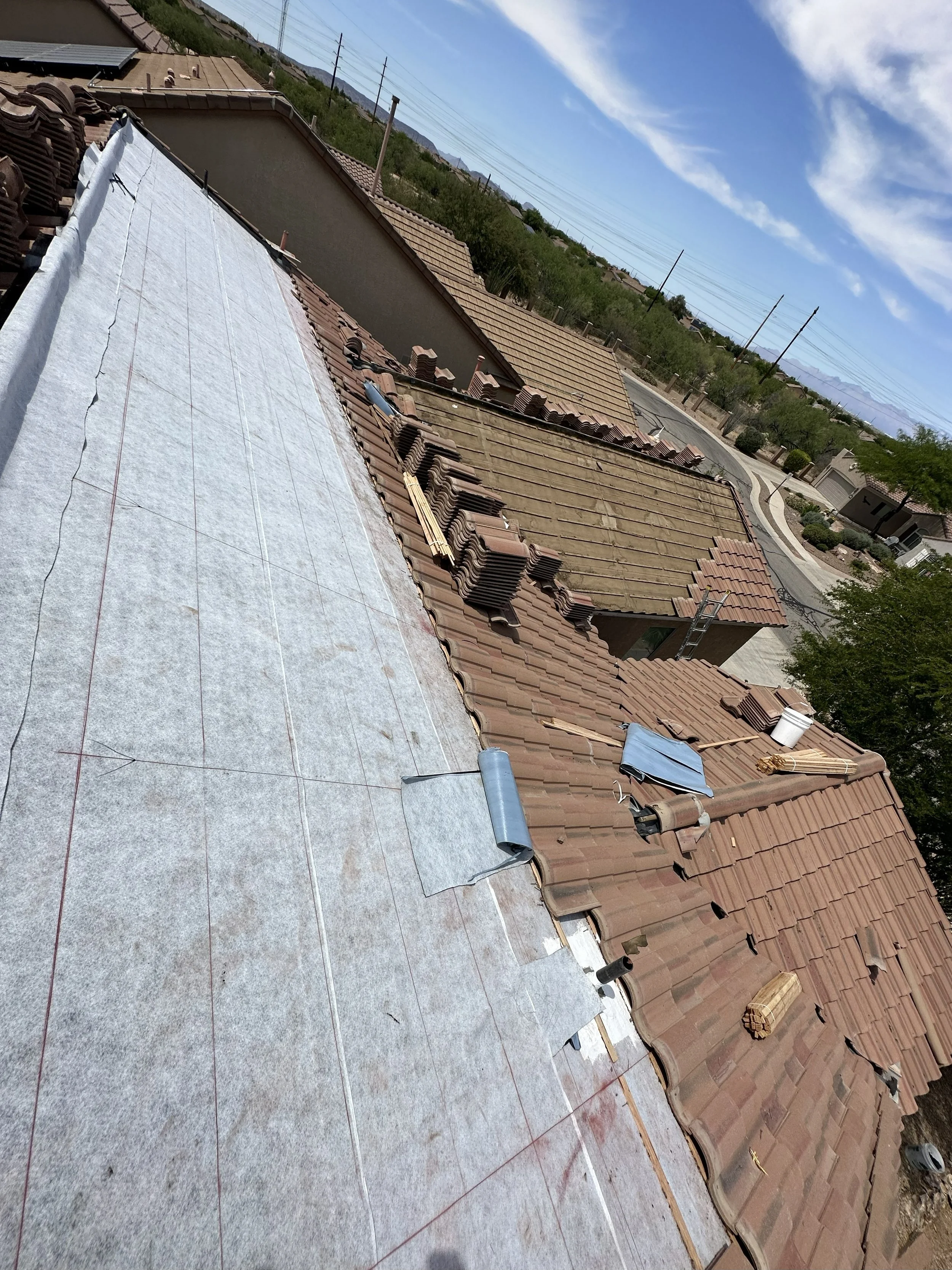 Roof under construction with felt paper and stacked tiles, partially completed with tools and building materials seen on top, in a residential neighborhood with other rooftops and a clear sky.