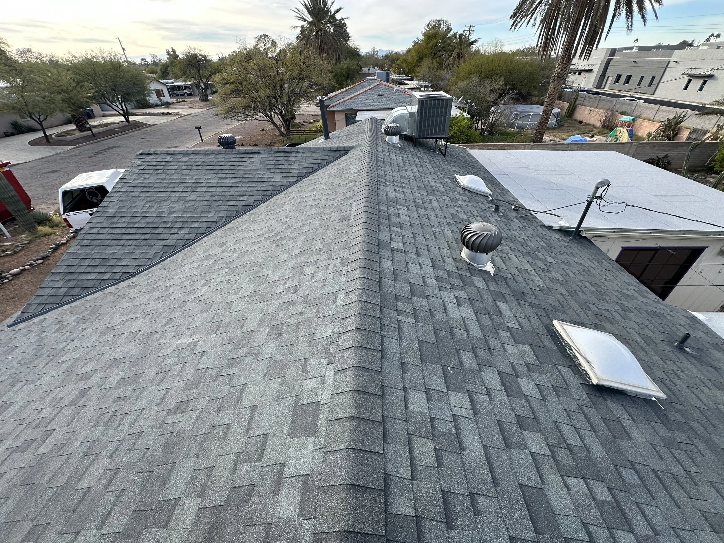 Aerial view of a rooftops featuring multiple vent pipes, skylights, and rooftop units in a residential area with trees and neighboring houses visible.
