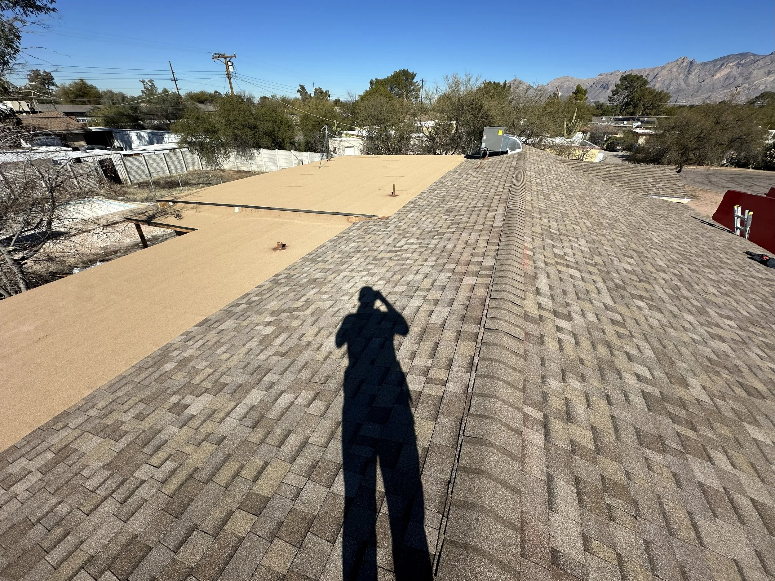 View of a rooftops with brown asphalt shingles and a small section of flat roof under construction, surrounded by trees, fences, and mountains in the distance under a clear blue sky.