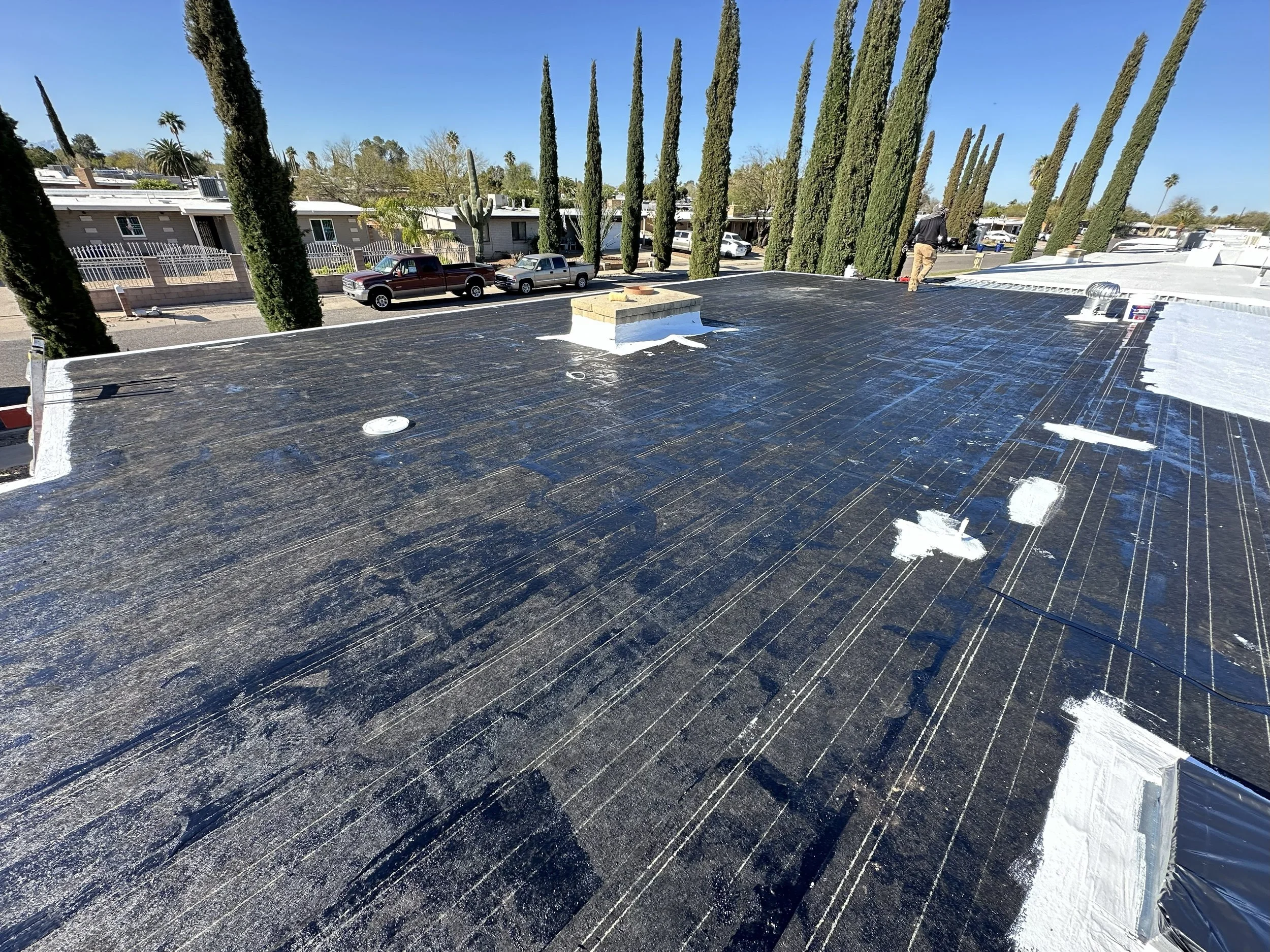 Flat roof under maintenance with black coating, man working in the background, surrounded by tall cypress trees and parked cars in a sunny residential neighborhood.