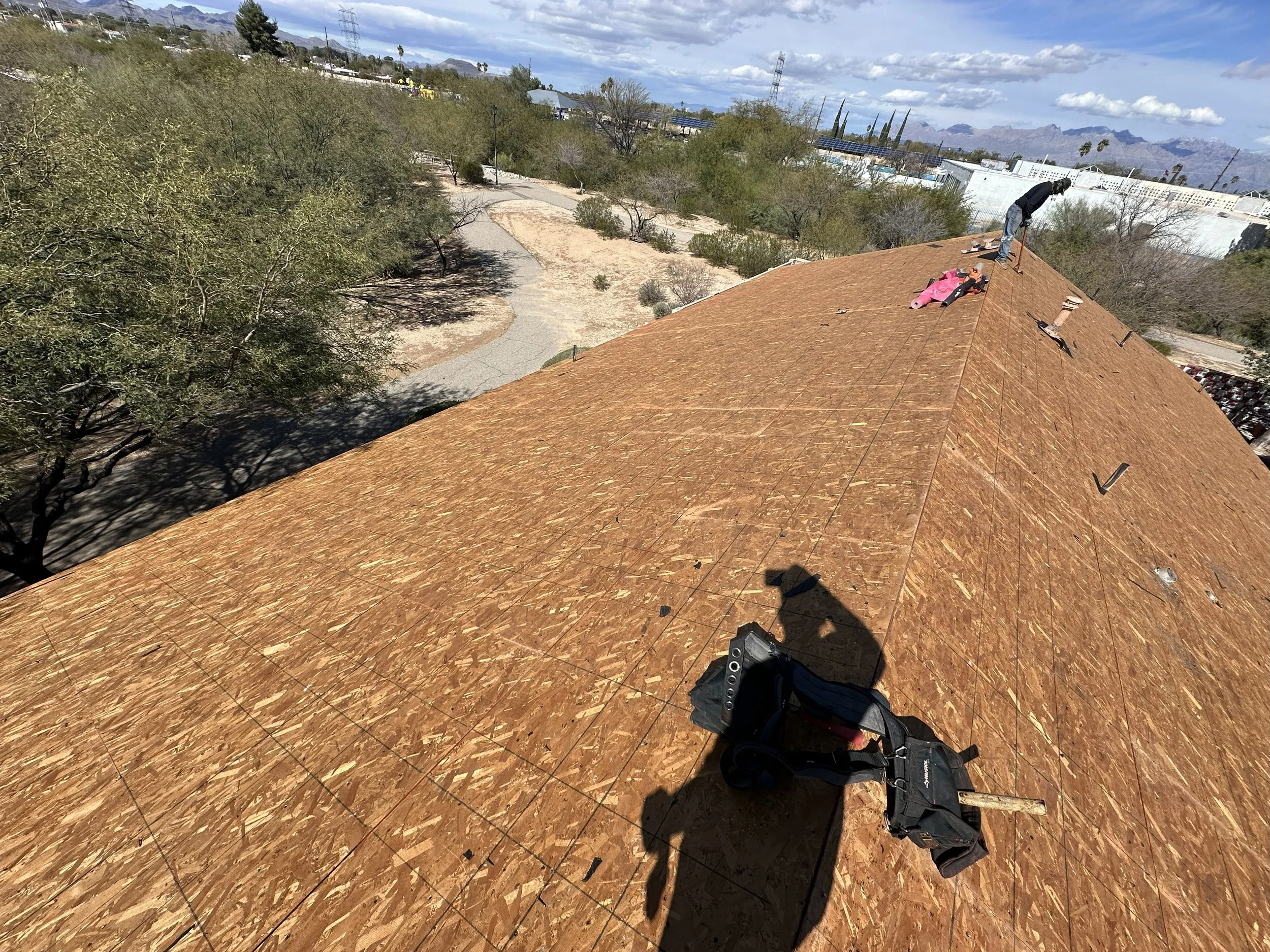A person is working on installing roofing on a building, with tools and materials on the roof. The view looks down from the roof showing a yard, trees, and a distant cityscape with mountains.