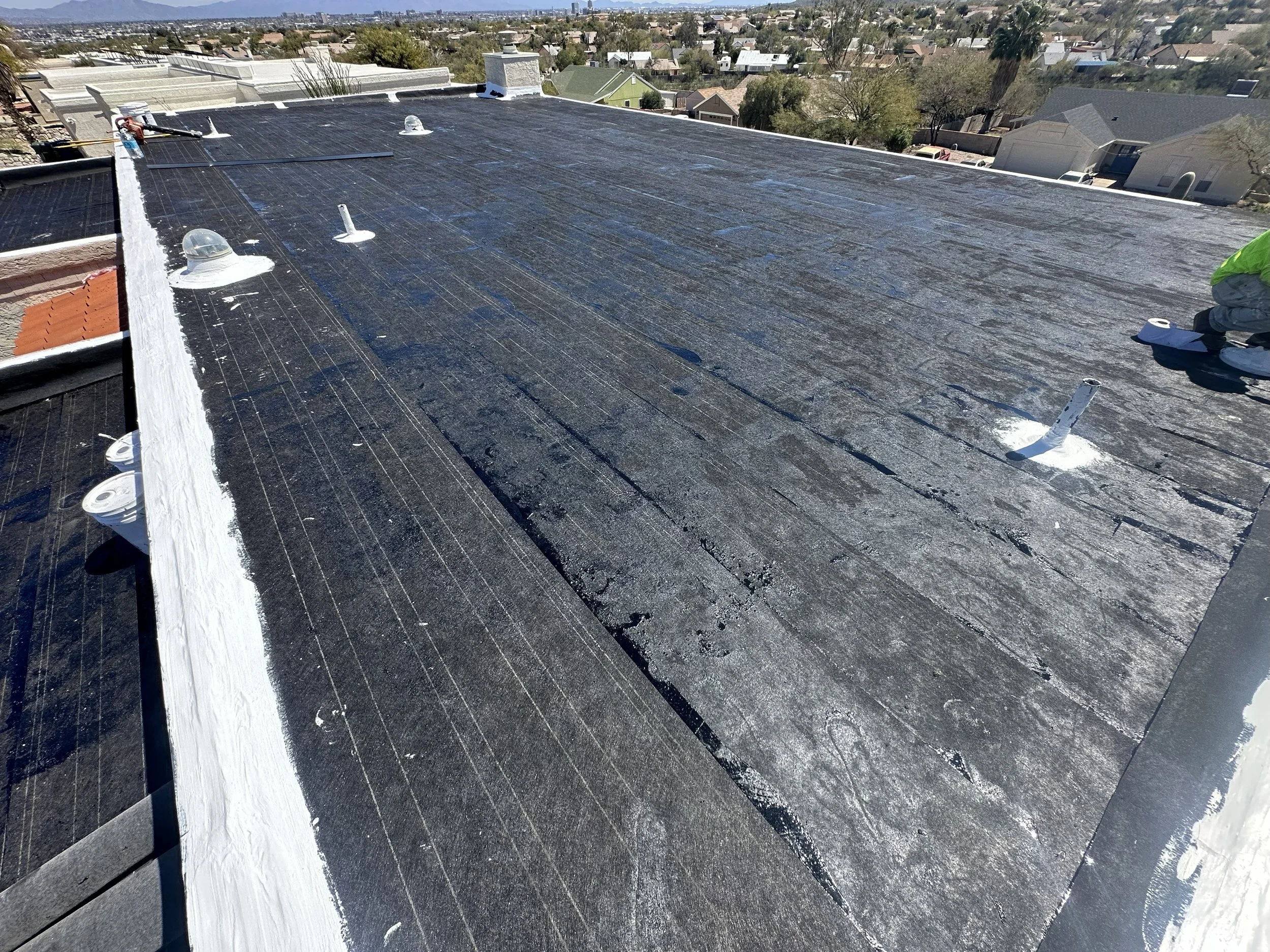 A residential rooftop under repair with new black roofing material installed, protective plastic sheeting around vents, and a worker in a green jacket on the right side. The view overlooks a neighborhood with trees and houses under a clear sky.