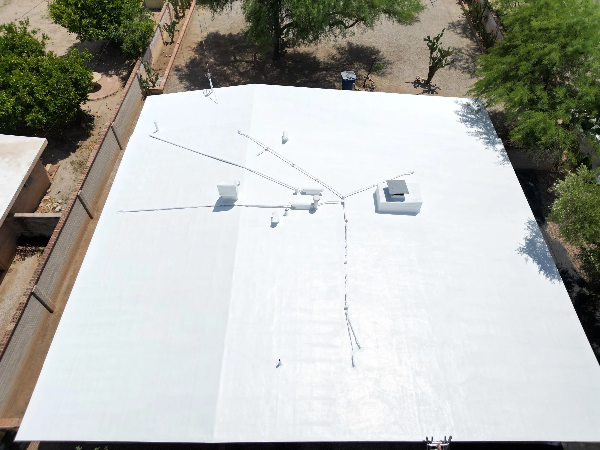 Top view of a white flat rooftop with several antennas, vents, and equipment, surrounded by trees and fences.