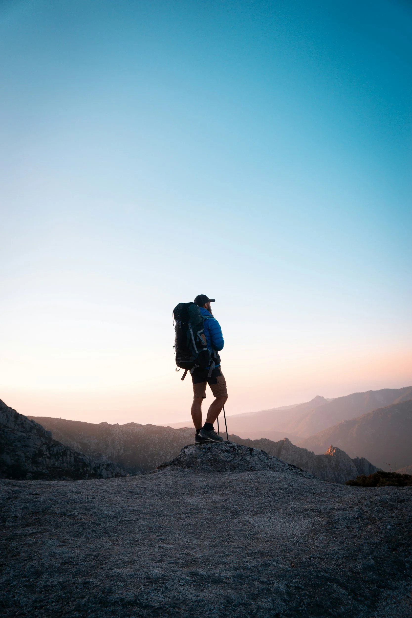 A hiker with a backpack and trekking poles standing on a large rock formation at sunrise or sunset, looking at distant mountains.