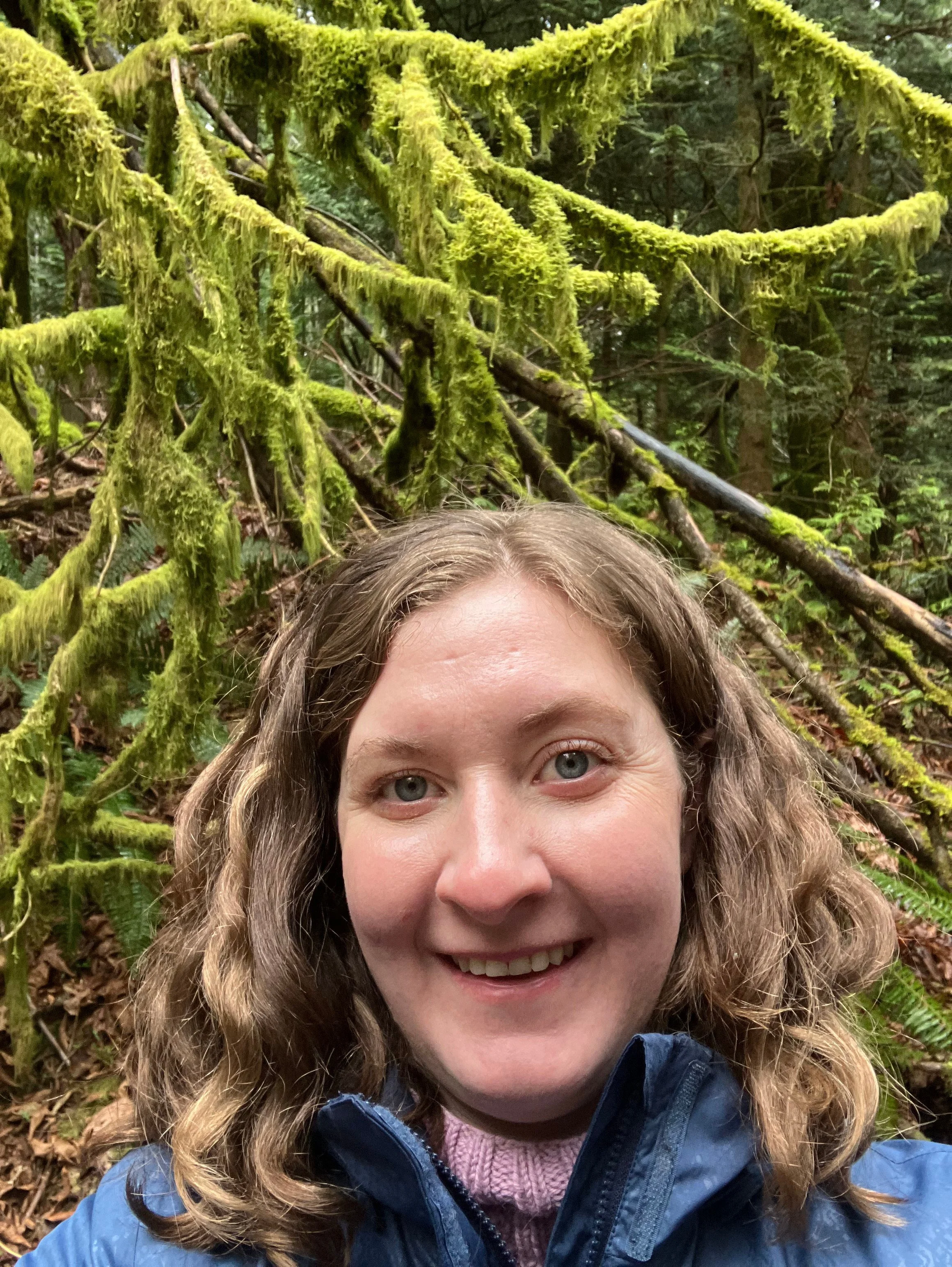 A woman with curly brown hair and a blue jacket taking a selfie in a lush, green forest with moss-covered branches in the background.