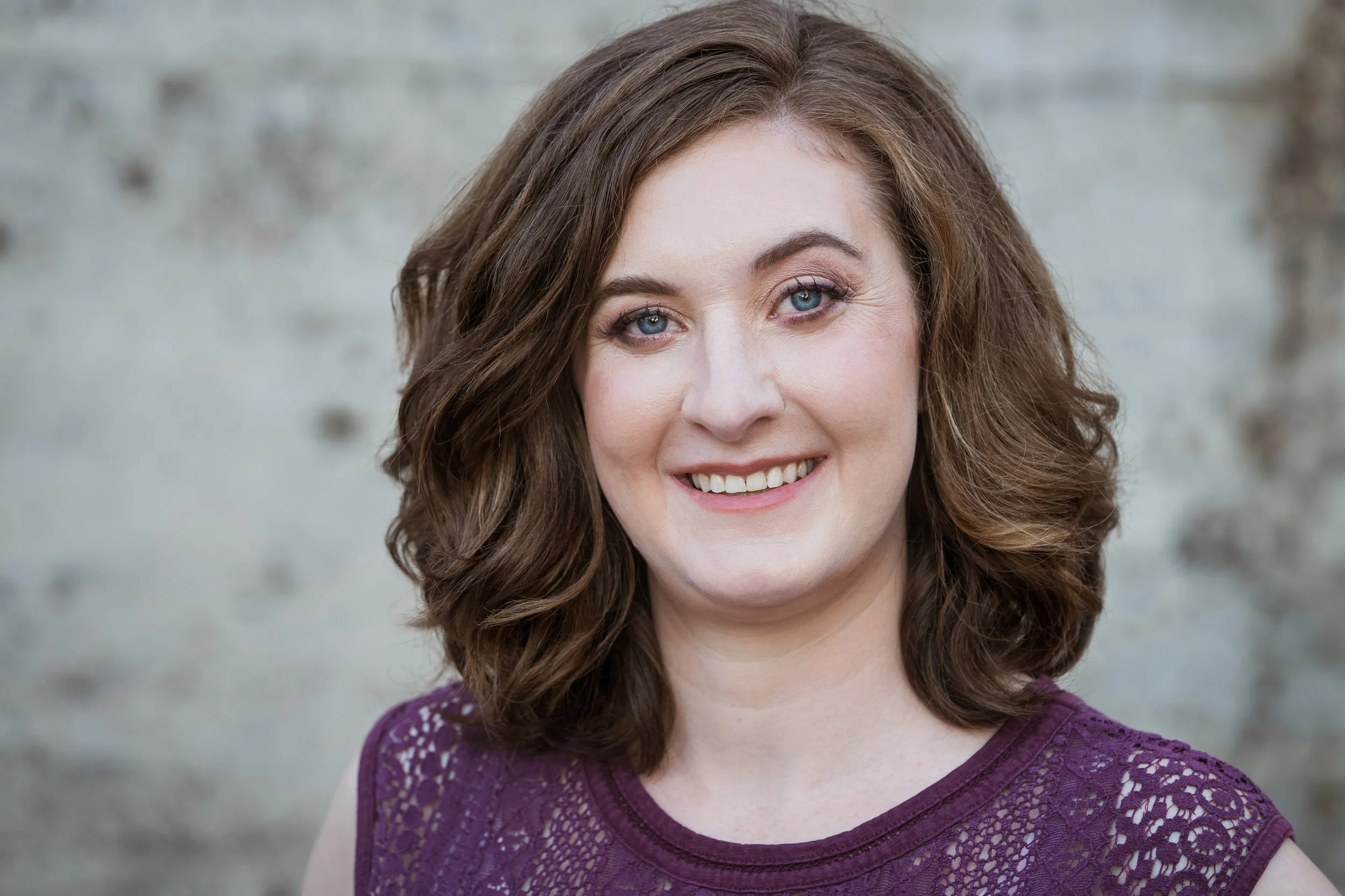 A smiling woman with shoulder-length curly brown hair, blue eyes, and wearing a purple lace top, standing in front of a blurred light gray background.