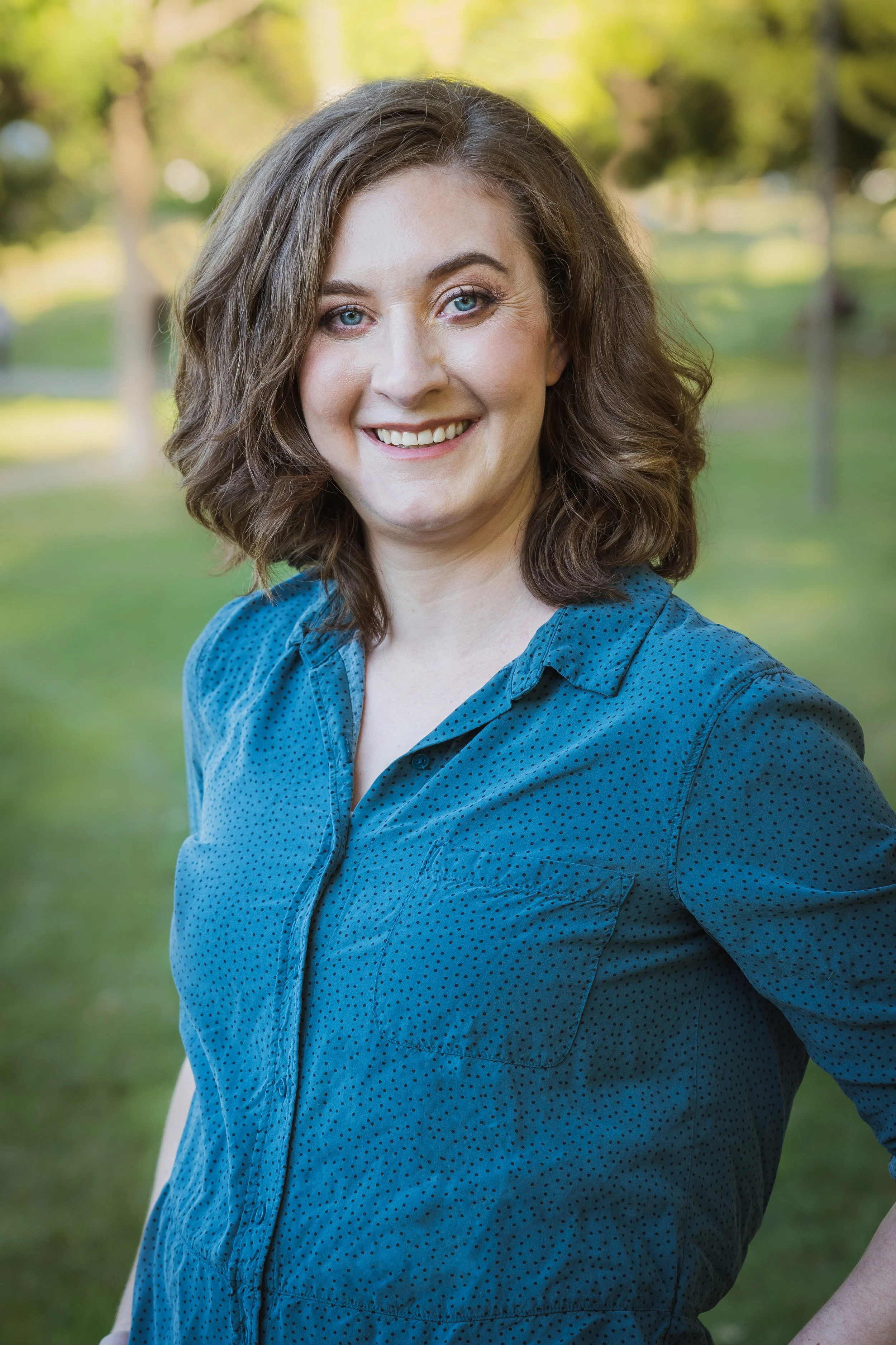 A woman with shoulder-length wavy brown hair and blue eyes, smiling in a park with green grass and trees in the background, wearing a blue, dotted button-up shirt.
