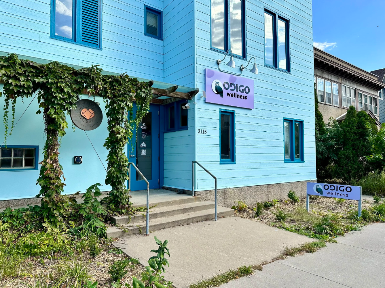 Blue building with signs for ODDGO Wellness, surrounded by small plants and bushes, with a sidewalk in front.