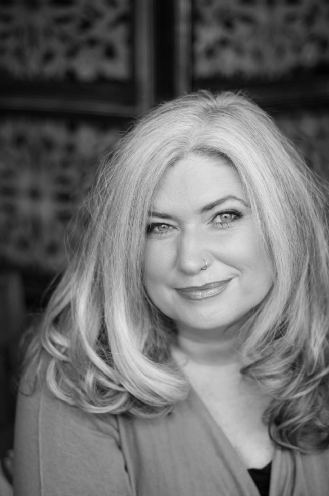 Black and white portrait of a woman with light-colored, wavy hair and a nose piercing, smiling gently, wearing a top with a rounded neckline.