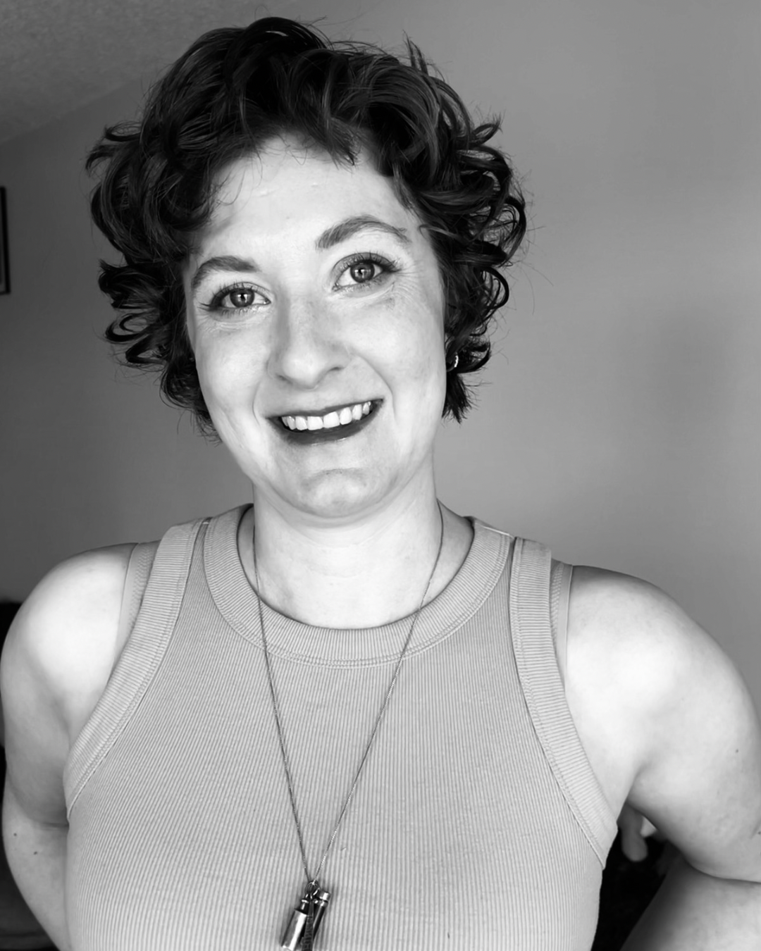 A smiling woman with curly hair wearing a sleeveless top and a necklace, posing indoors.