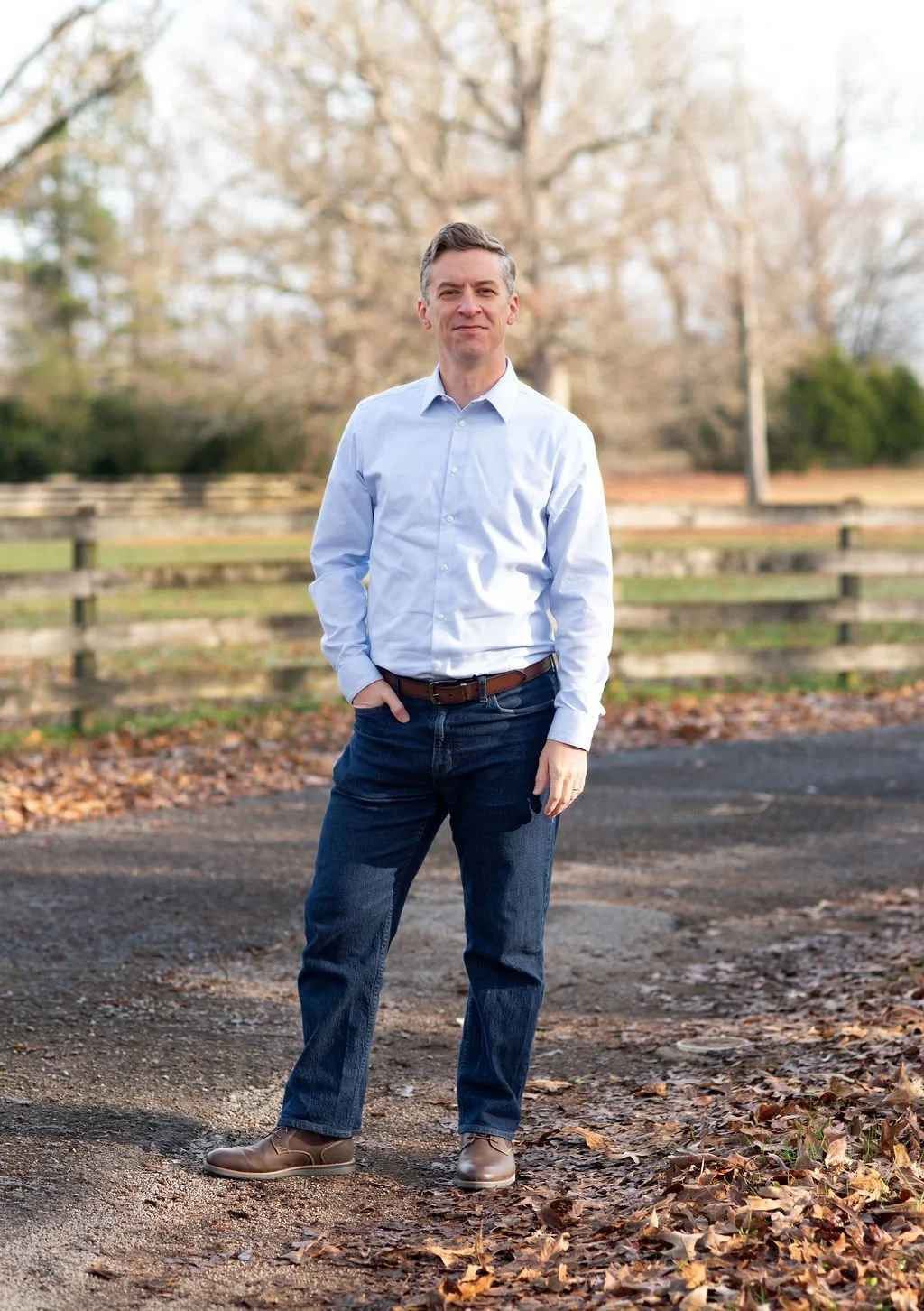 Michael Roberts stands outdoors on a dirt path with fallen leaves, wearing a light blue dress shirt, dark jeans, and brown shoes, with a wooden fence and leafless trees in the background.