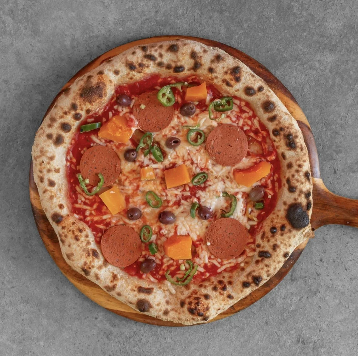 A pepperoni pizza with green peppers, black olives, and chunks of orange bell peppers on a wooden pizza board.