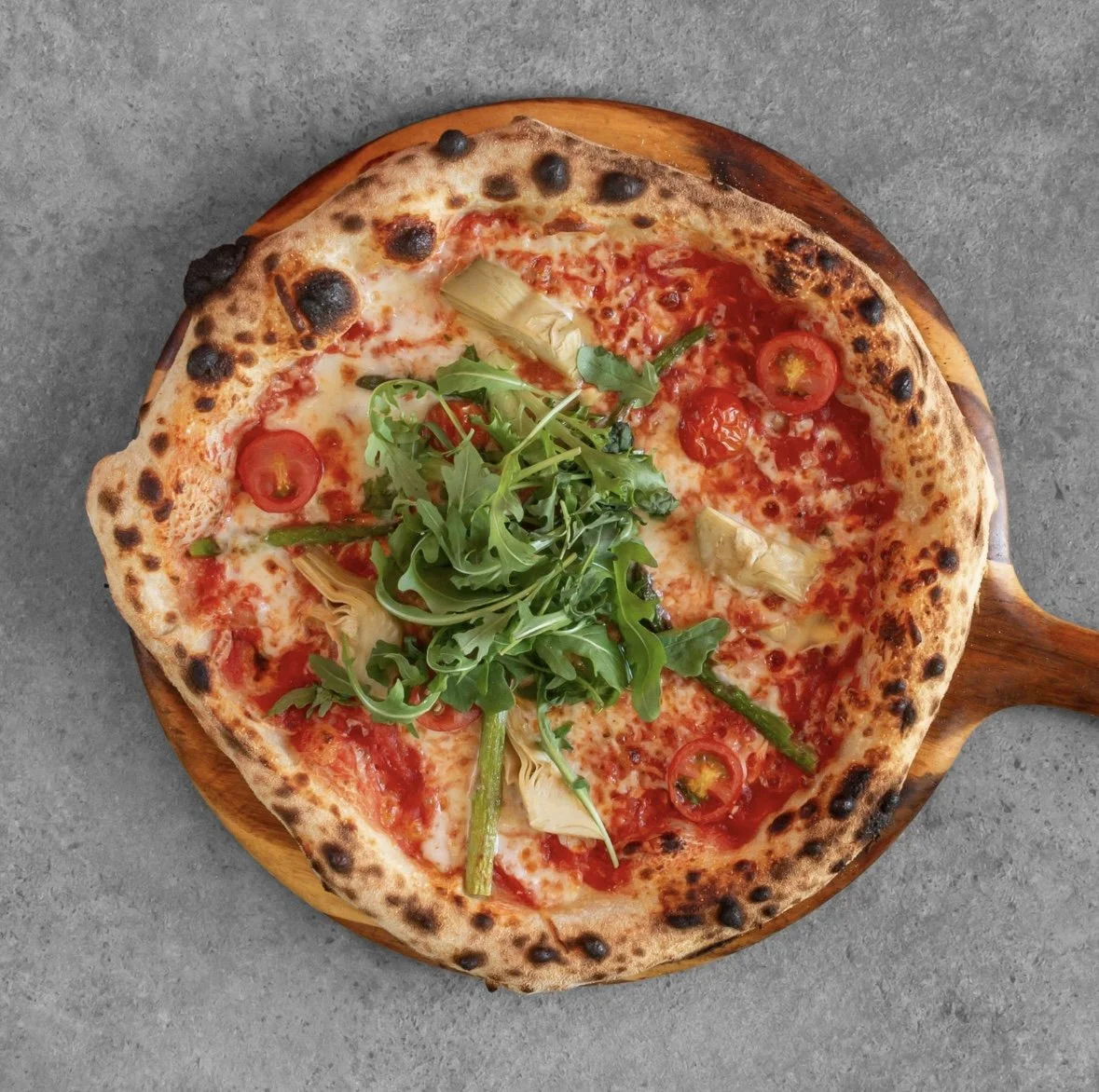 Fresh pizza with cherry tomatoes, arugula, artichoke hearts, and green peppers on a wooden serving board.
