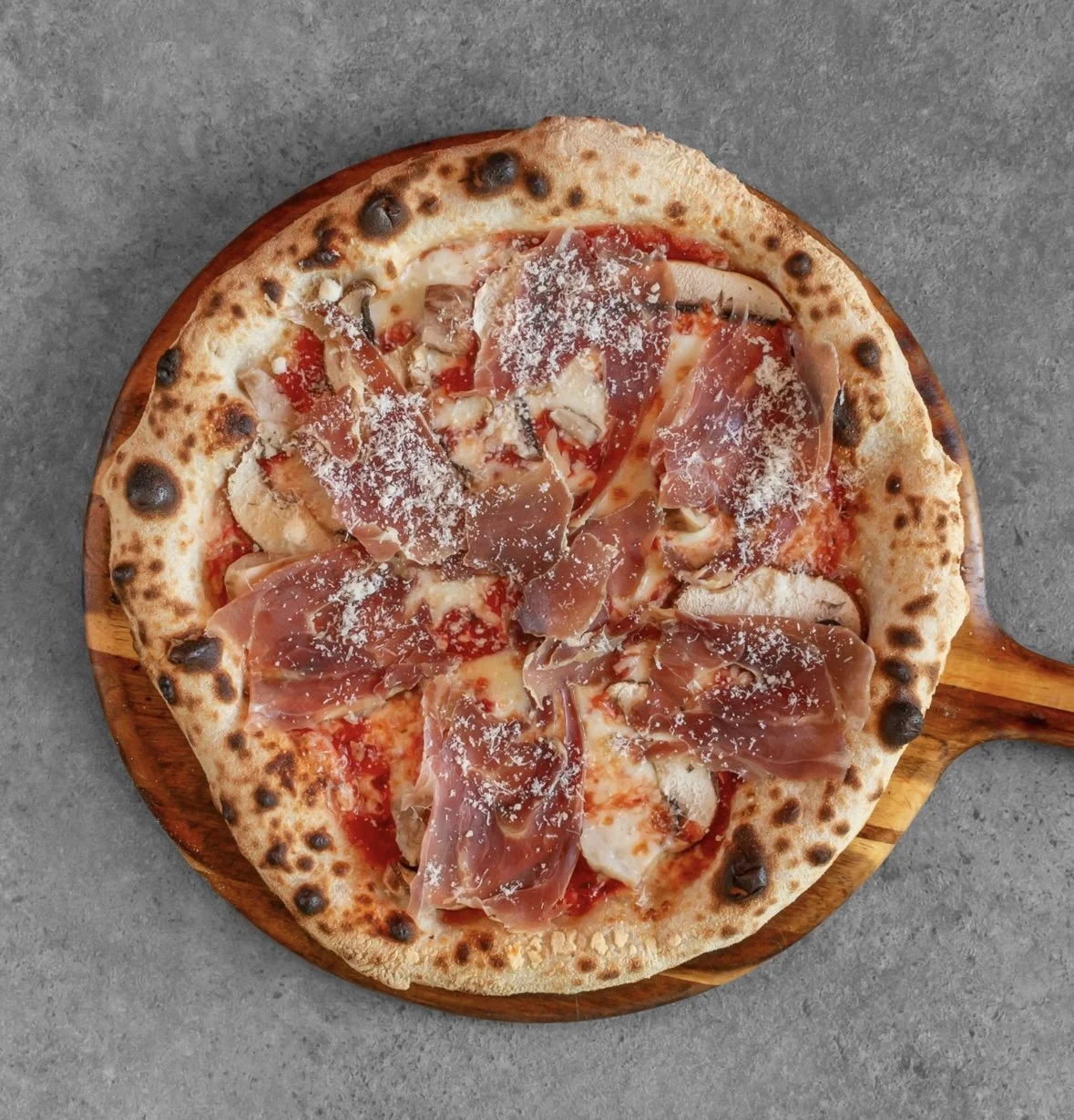 Top view of a prosciutto mushroom pizza with cheese on a wooden pizza board, placed on a gray surface.