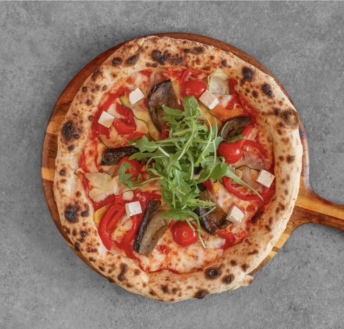 A pizza on a wooden tray with toppings including cherry tomatoes, red bell peppers, grilled eggplant, arugula, and small cubes of cheese, set on a gray surface.