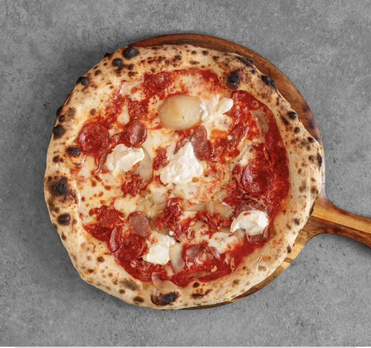 A pizza with pepperoni, white cheese, and a whole garlic clove on top, served on a wooden pizza board on a gray surface.