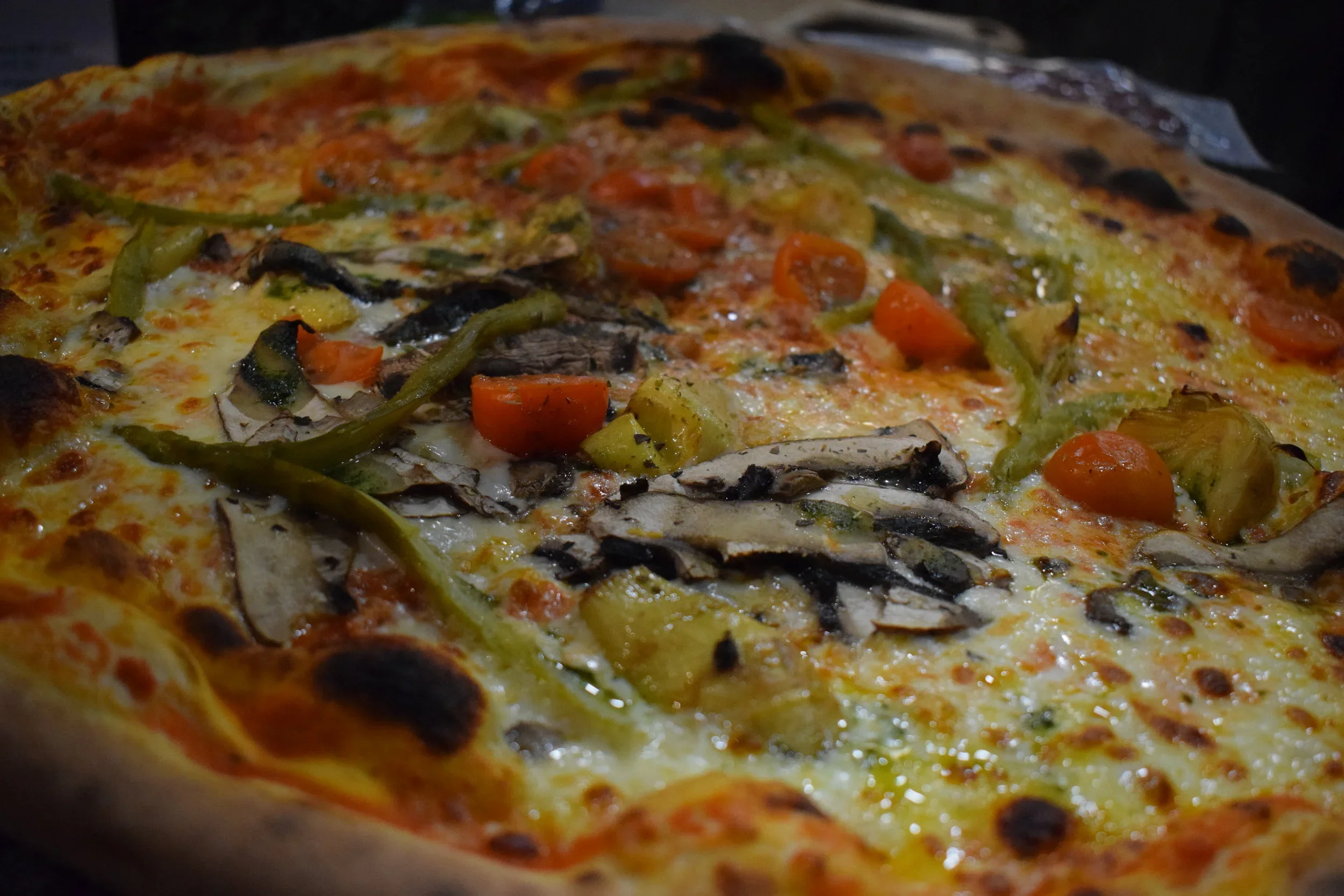 Close-up of a sliced pizza with cheese, mushrooms, cherry tomatoes, green peppers, and pineapple on a wooden surface.