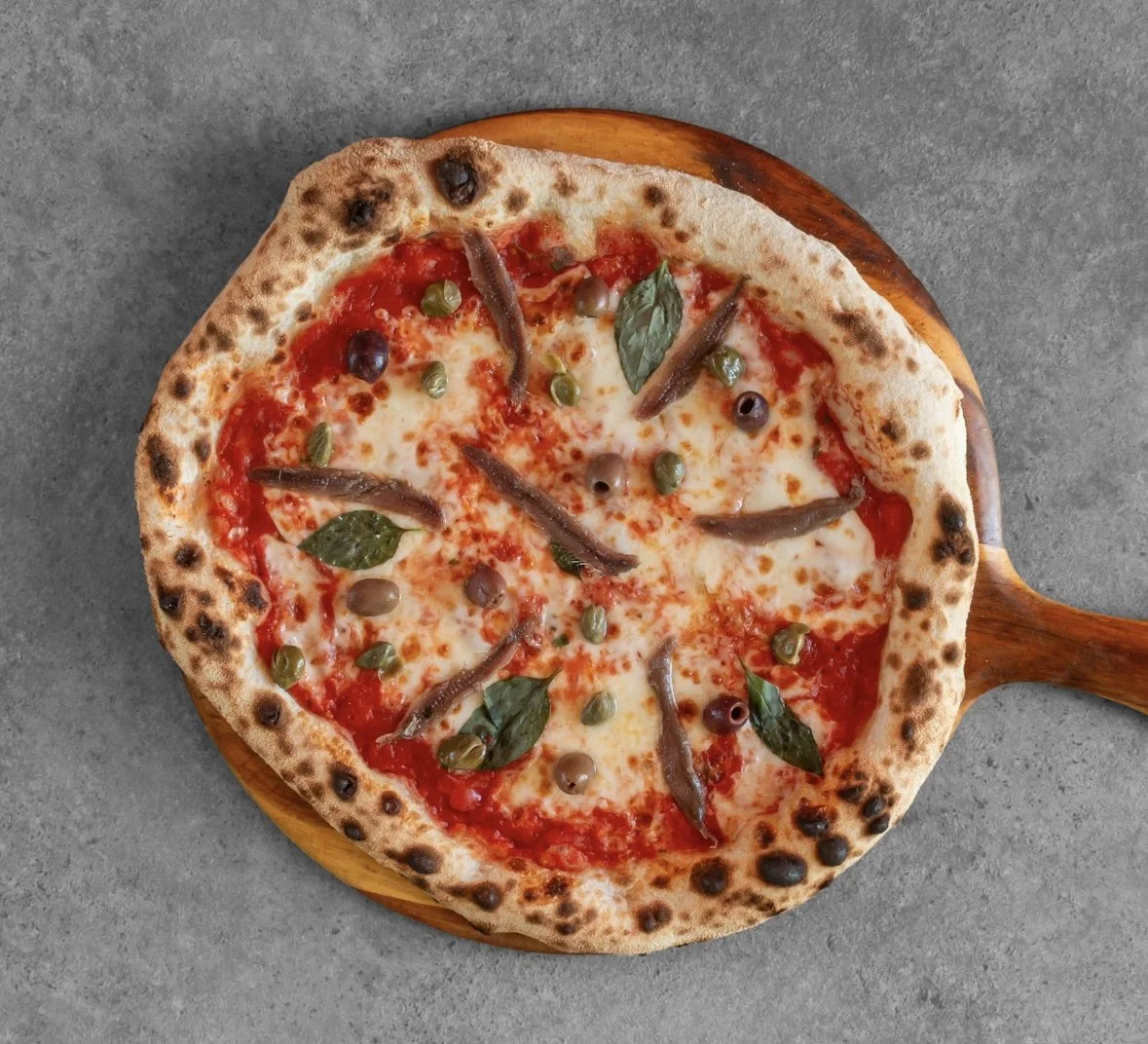 A Neapolitan-style pizza with tomato sauce, melted cheese, anchovies, capers, and basil leaves on a wooden serving tray.