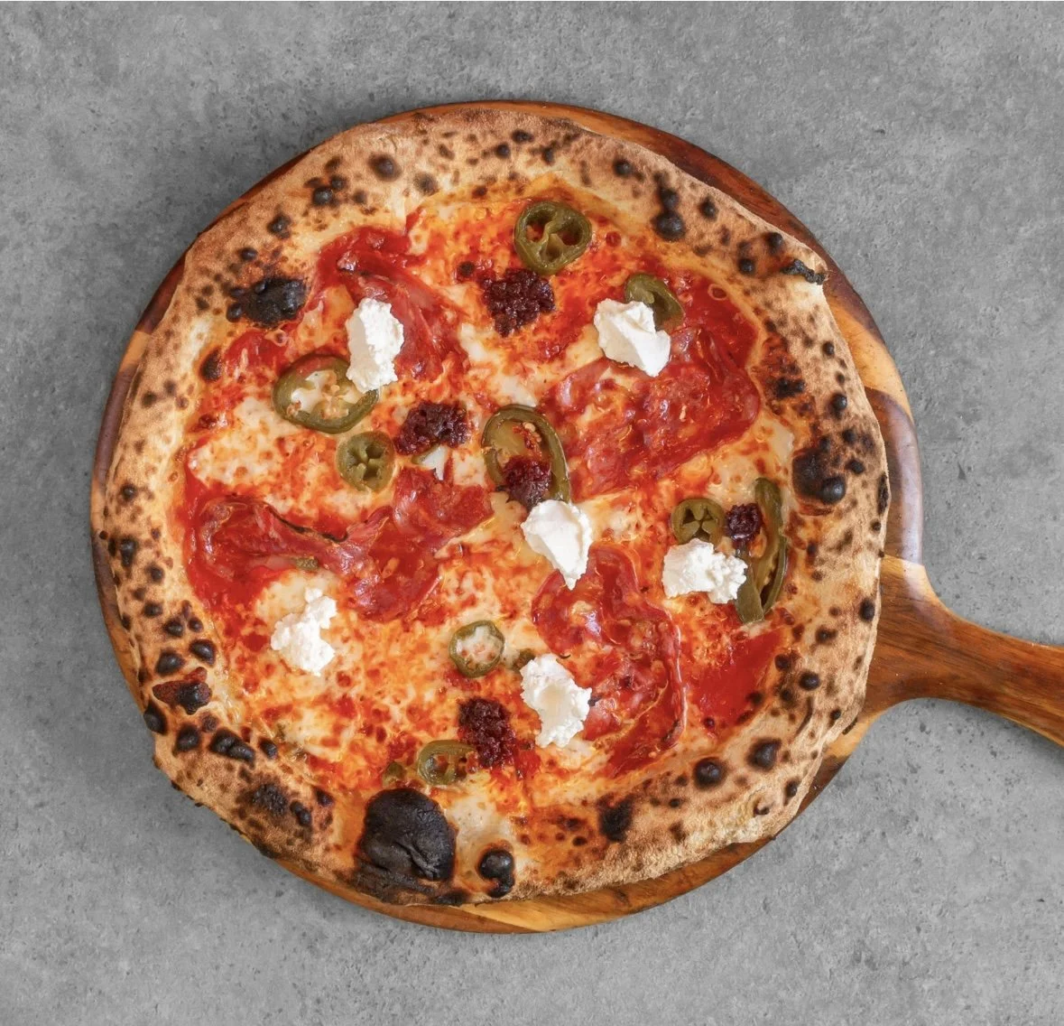 Neapolitan-style pizza with jalapeños, dollops of white cheese, and tomato sauce on a wooden pizza peel.