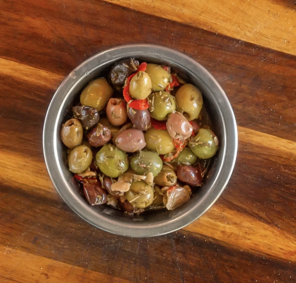 A bowl of assorted green and black olives with red pepper flakes on a wooden surface.