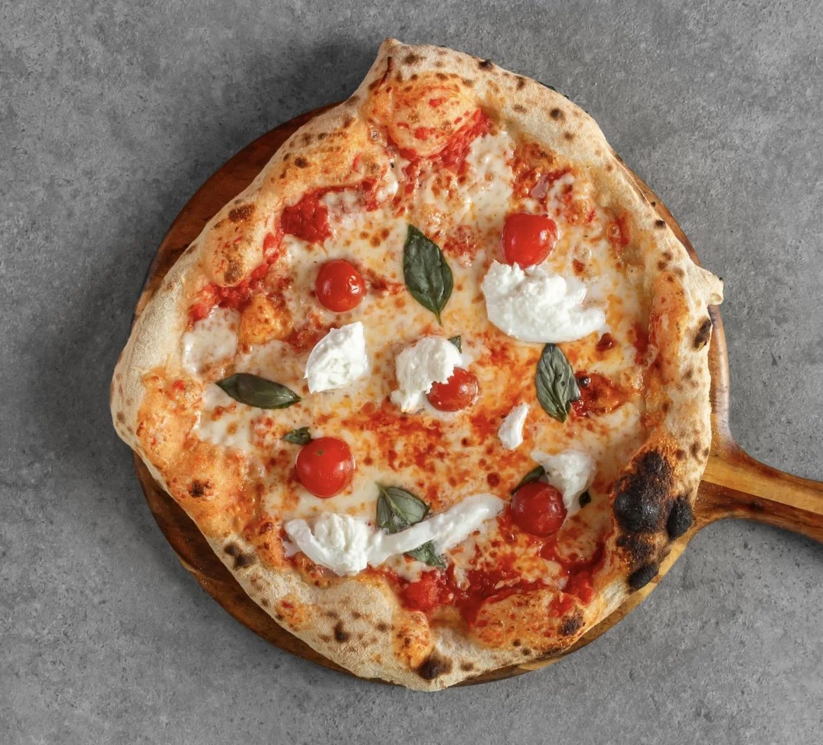 Slice of Margherita pizza with cherry tomatoes, basil, and mozzarella on a wooden pizza paddle.