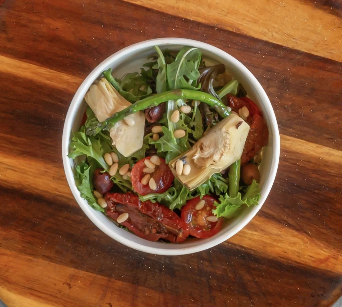 A bowl of mixed salad with artichoke, cherry tomatoes, greens, sunflowers seeds, and roasted red peppers on a wooden surface.